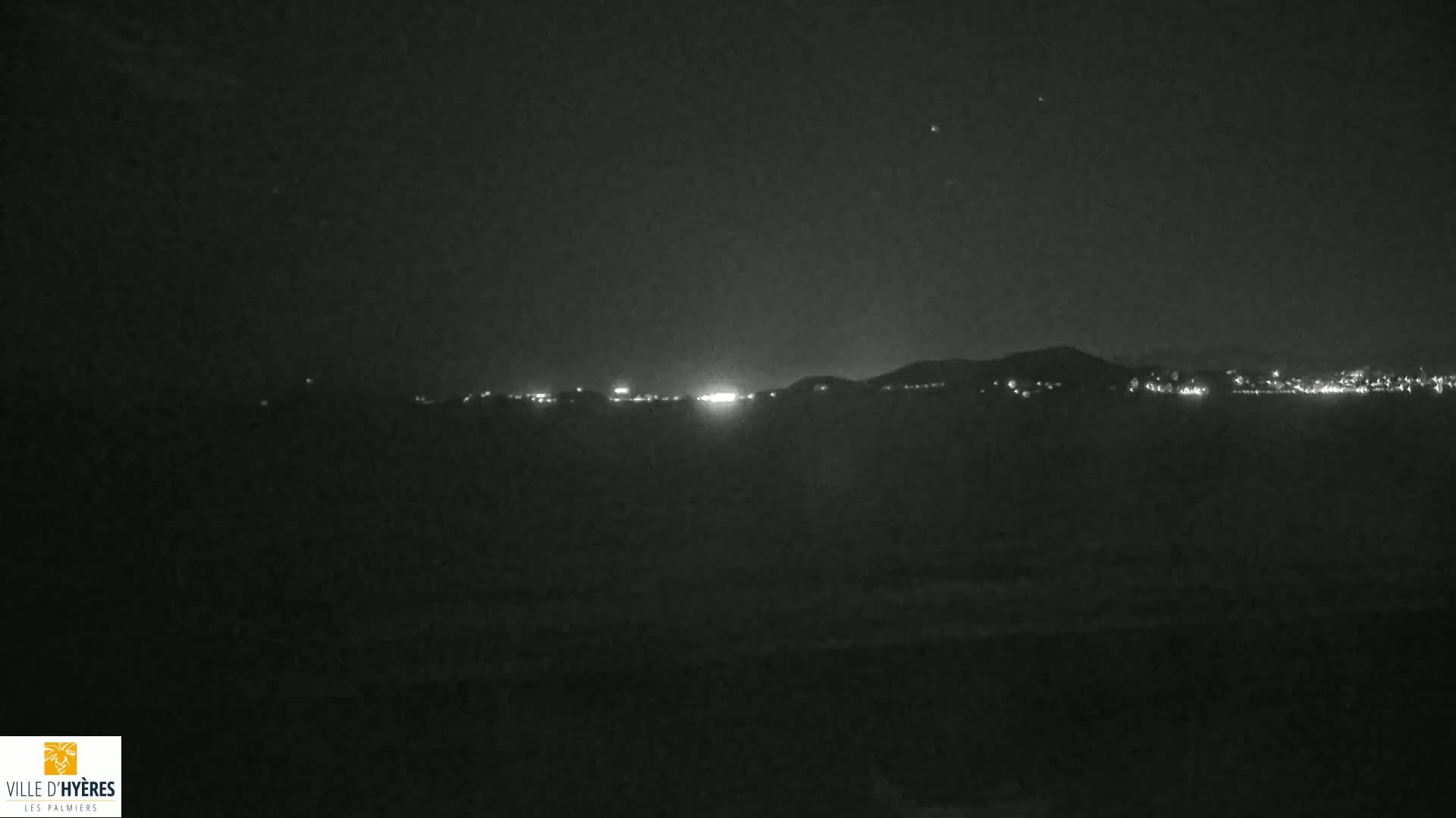 A nighttime view shows a dark, low-lit shoreline with a distant city and hills illuminated by lights under a clear, starry sky.