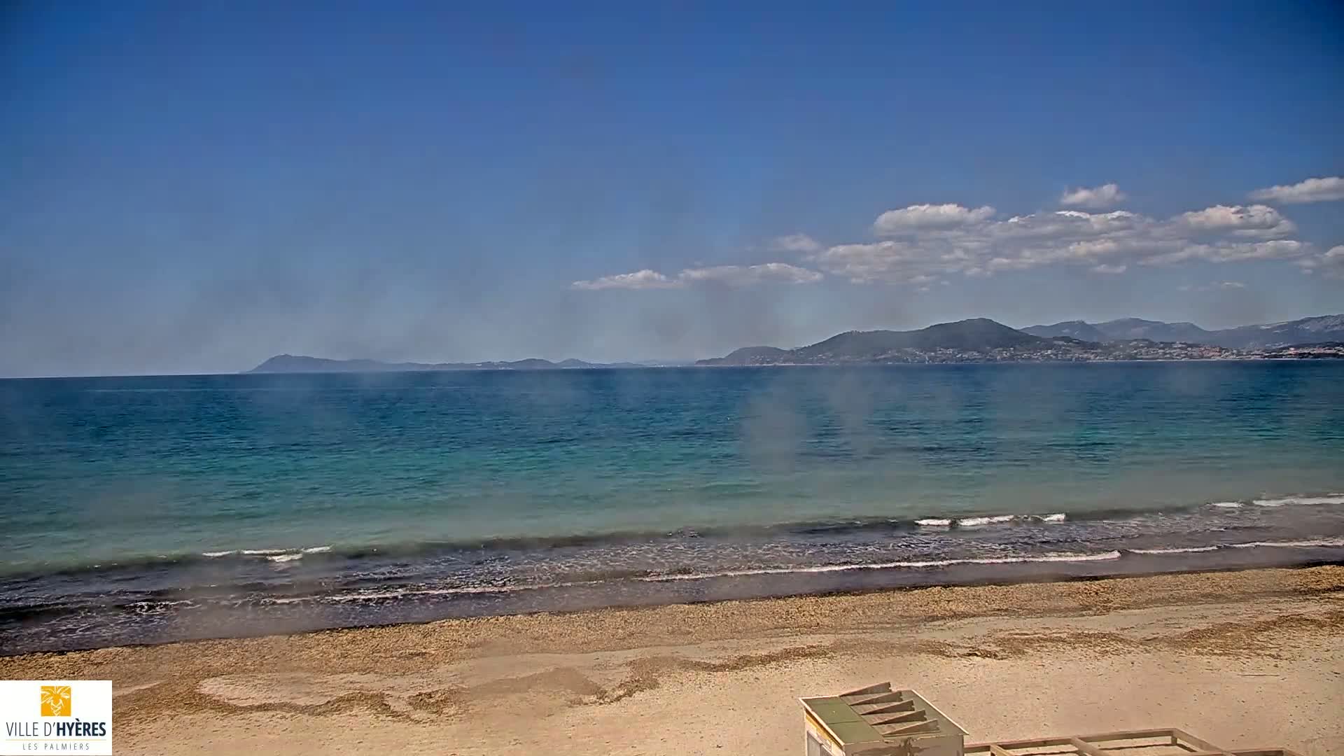A sandy beach with gentle waves laps the shore of a calm, turquoise sea under a mostly sunny sky, with distant mountains visible on the horizon.