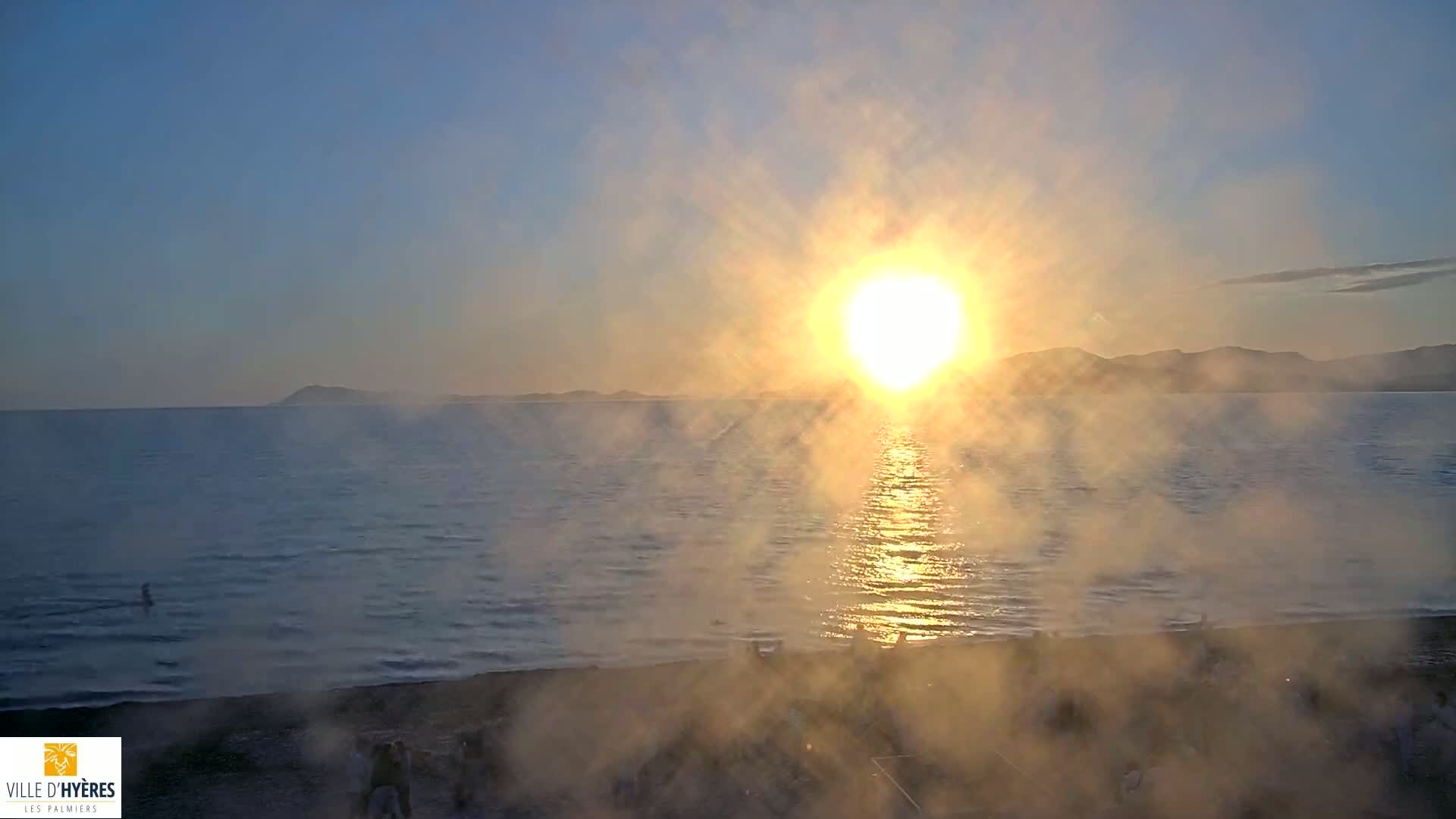A hazy sunrise over a calm sea with a beach in the foreground, partially obscured by mist or fog.
