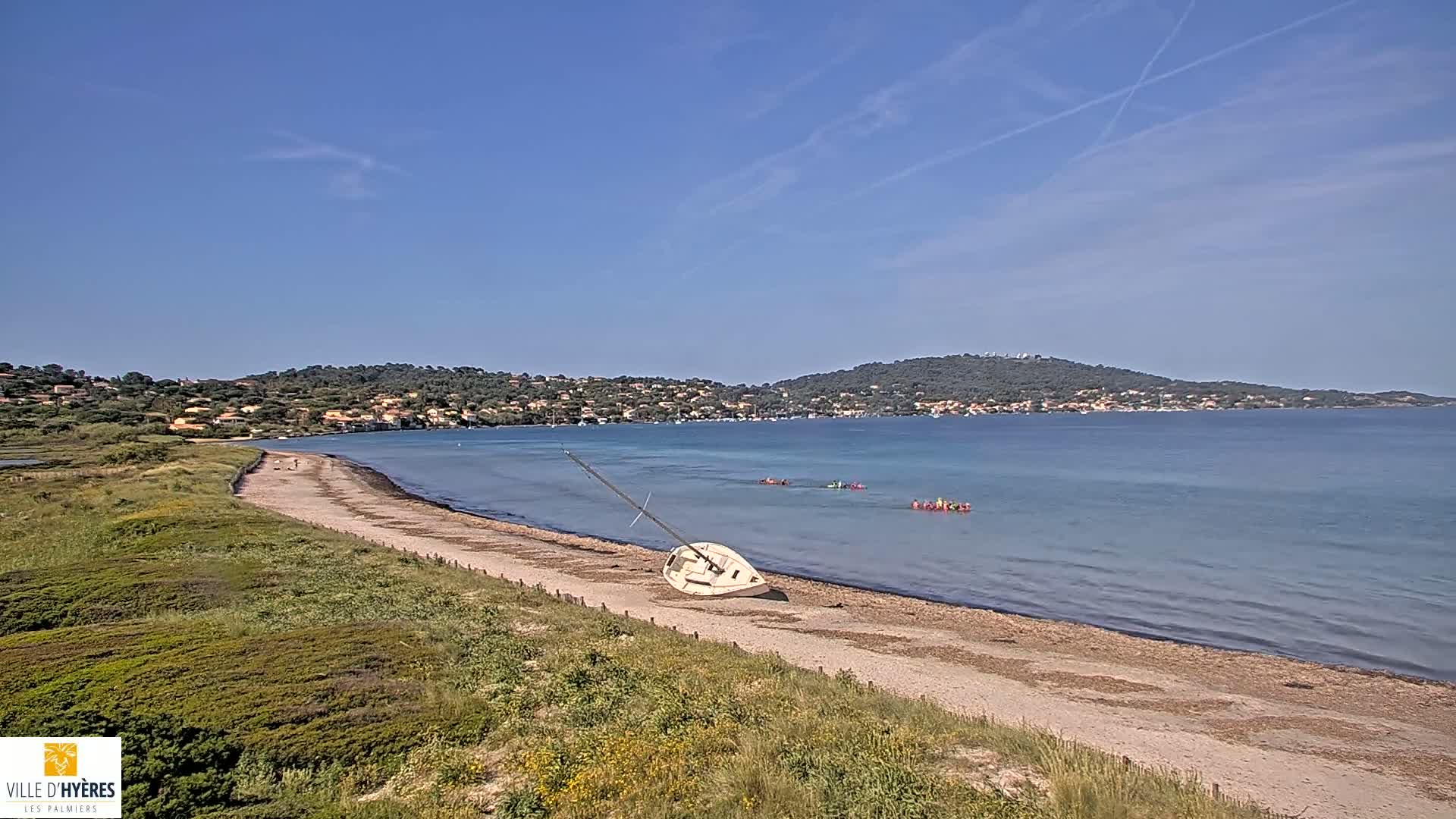 A sandy beach with a capsized sailboat lies on the shore, under a clear blue sky, with calm water and people in the distance kayaking near a hilly coastline.