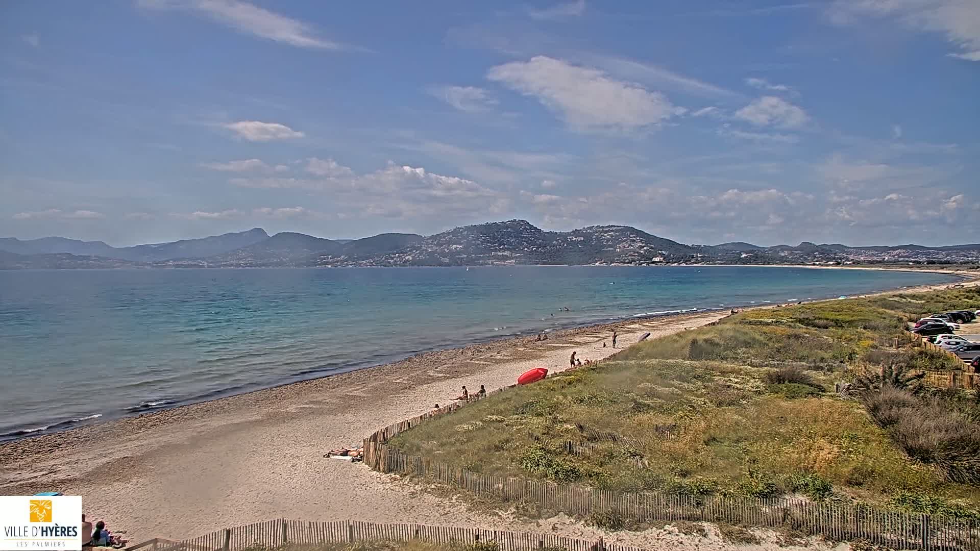 A sandy beach with scattered people stretches along a calm, turquoise sea under a partly cloudy sky, with mountains visible in the distance.