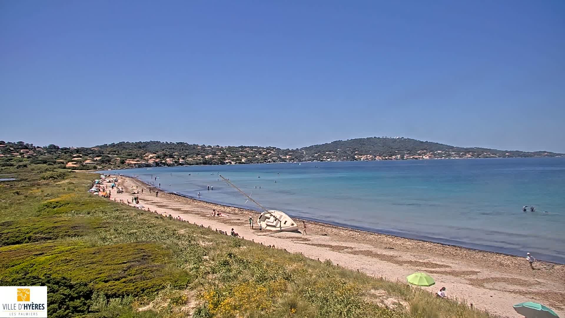 A sandy beach with a beached sailboat is populated with people on a sunny day, overlooking a calm, turquoise bay and distant hills.