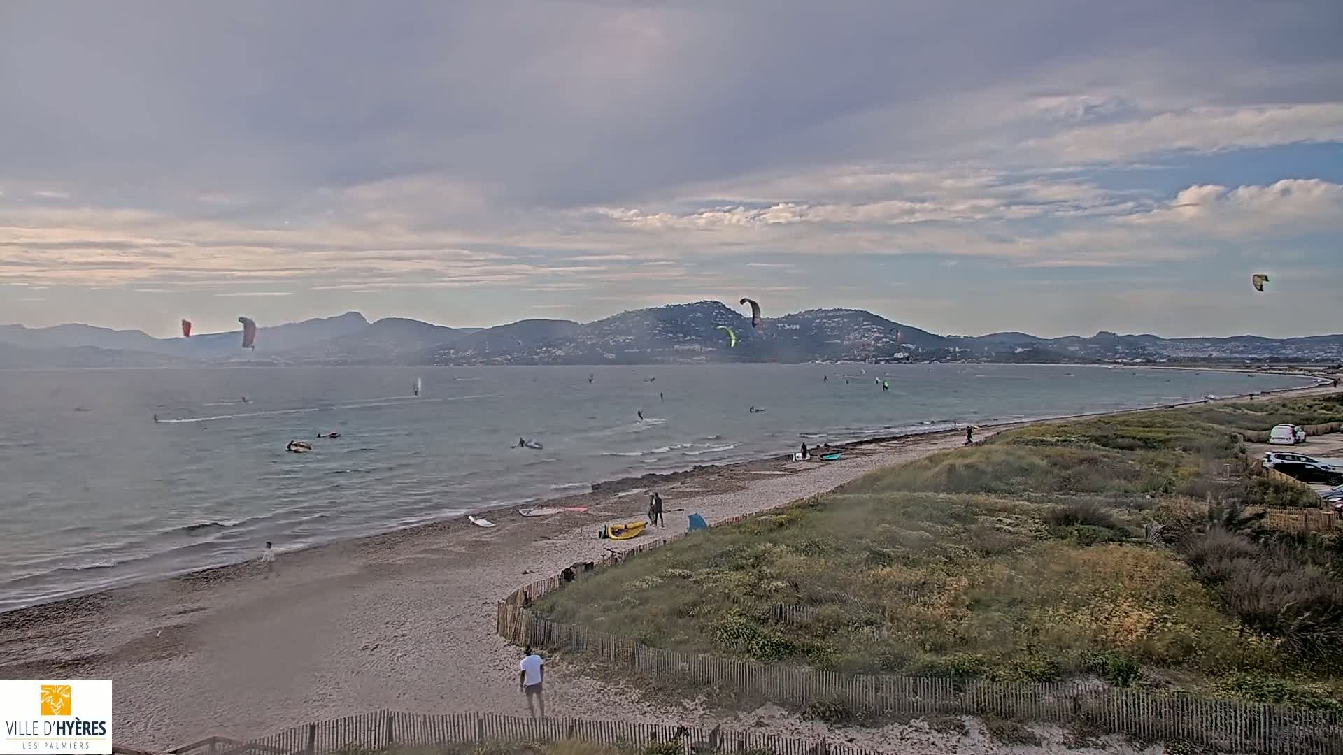 A sandy beach with several kite surfers in the water, under a partly cloudy sky, with mountains in the distance.