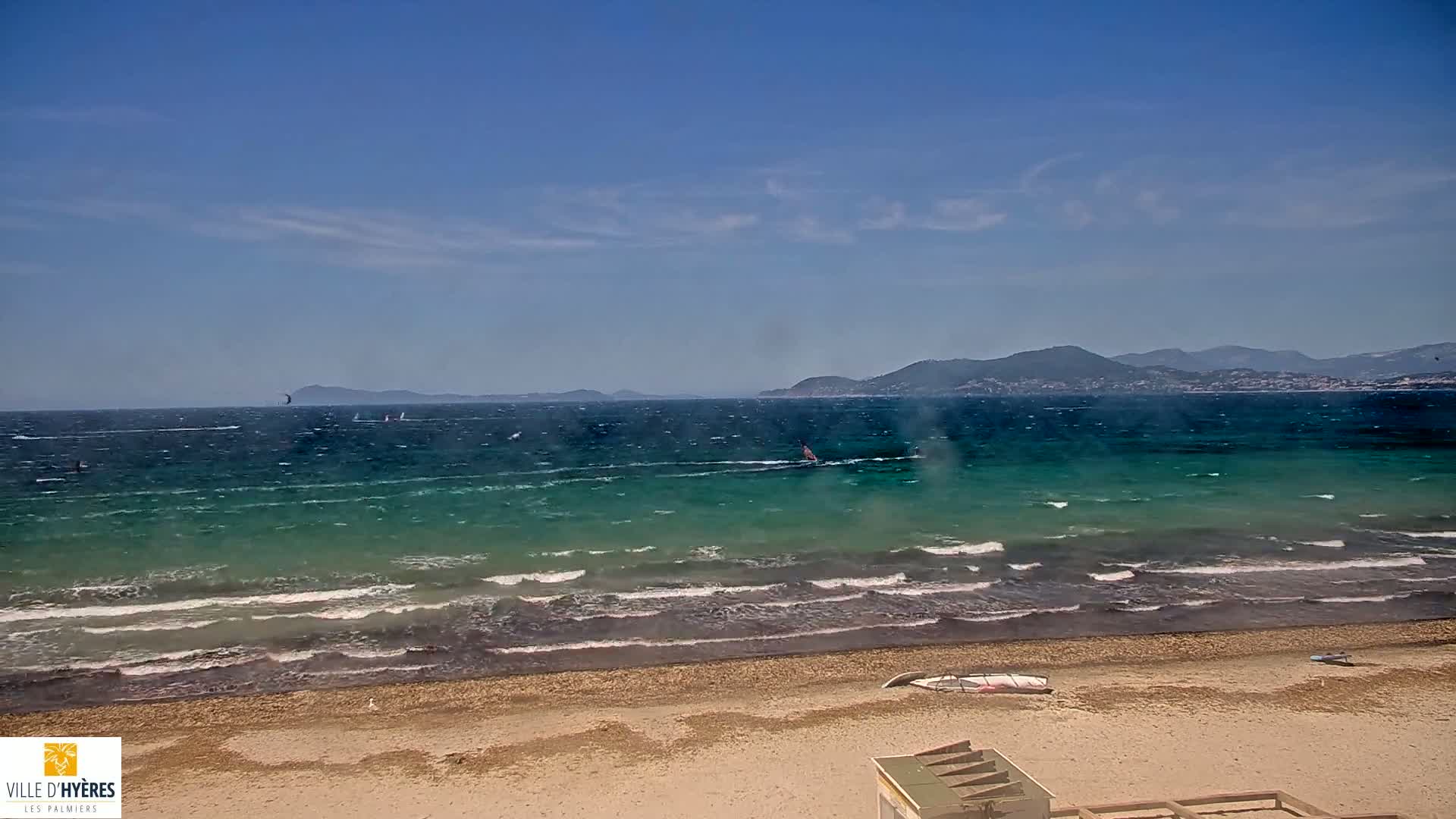 A windy day at a sandy beach shows a teal-colored sea with small waves and several windsurfers, with distant mountains visible on the horizon under a mostly clear blue sky.