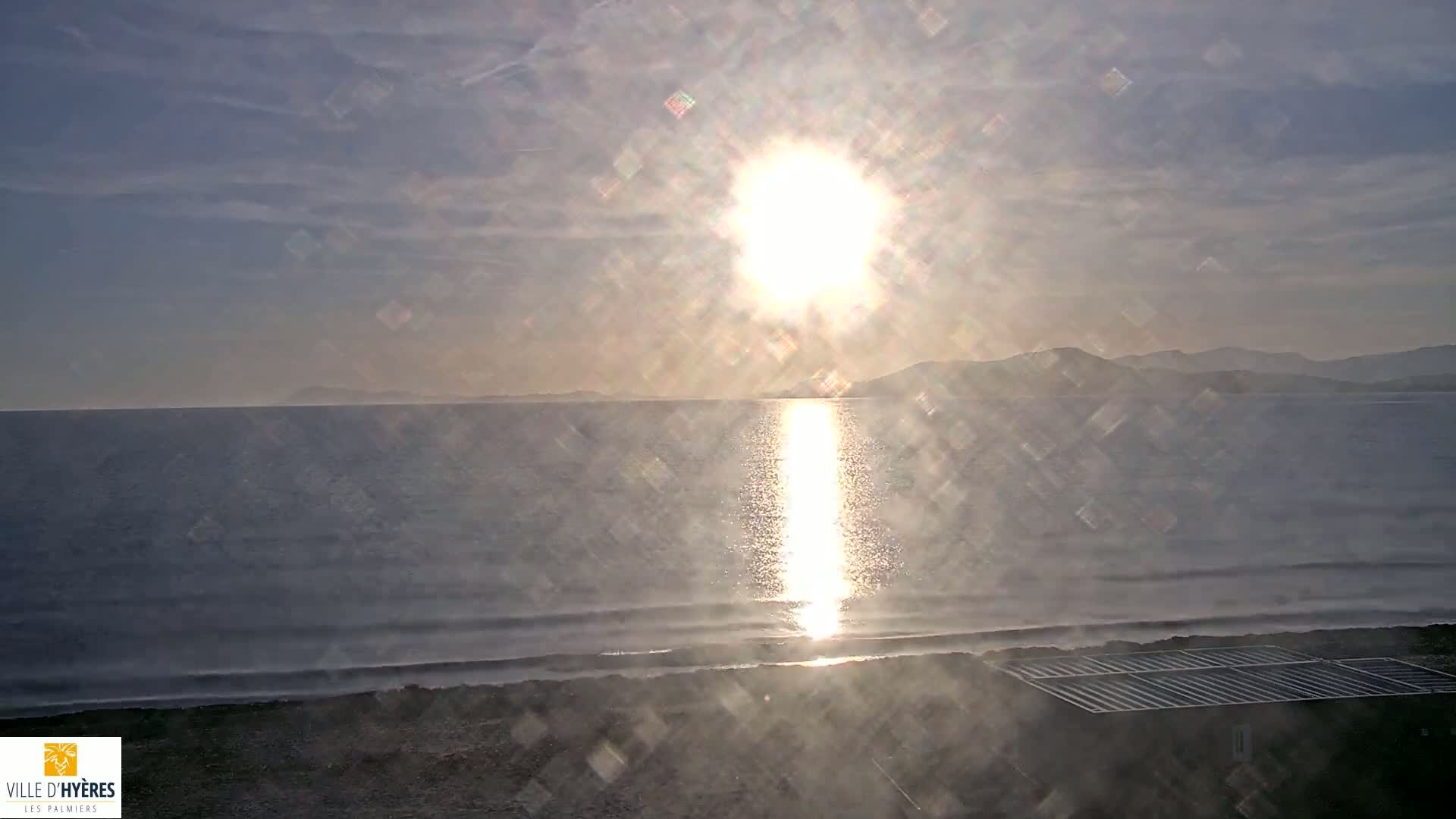 A bright sun shines over a calm sea with a distant shoreline and mountains under a partly cloudy sky.