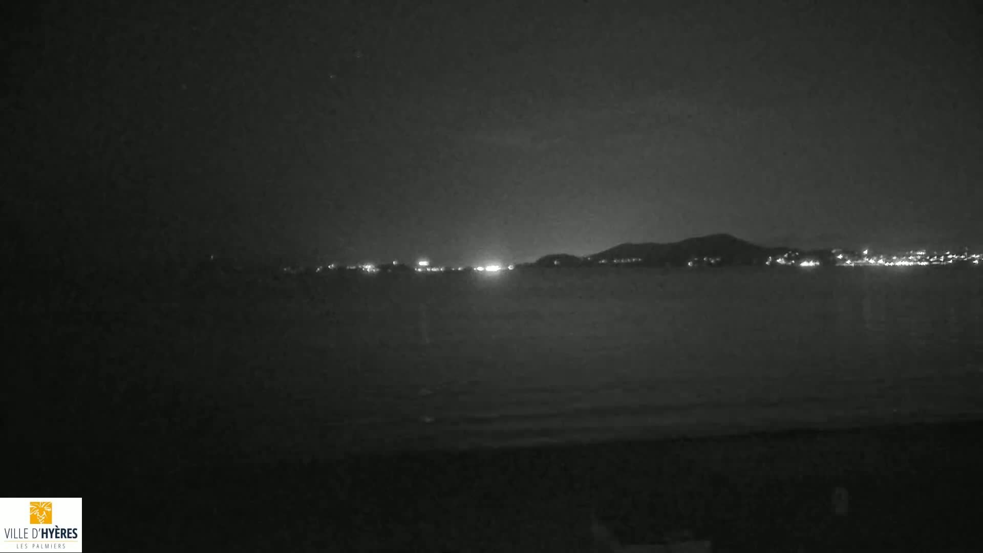 A nighttime view of a calm body of water with a distant, dimly lit shoreline and hills under a dark sky.