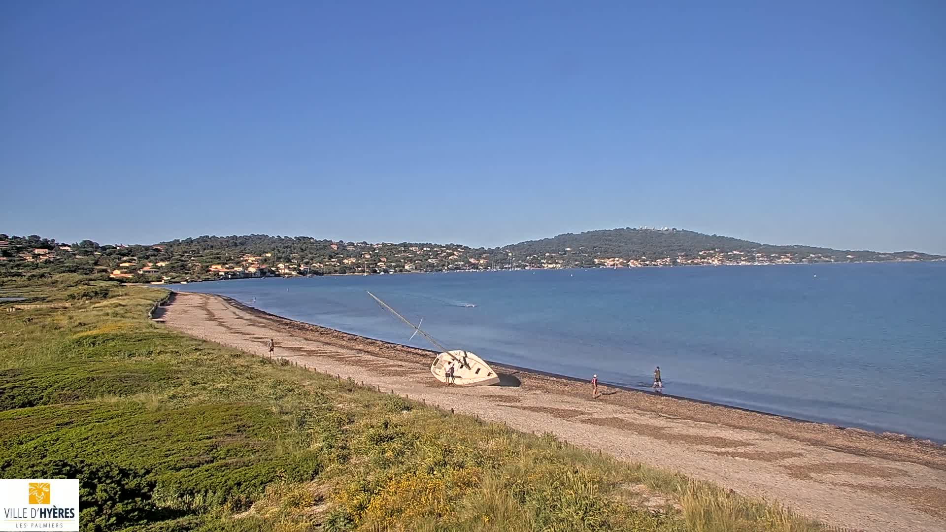 A sailboat lies on its side on a sandy beach next to calm, clear blue water under a clear blue sky, with a town visible in the distance.
