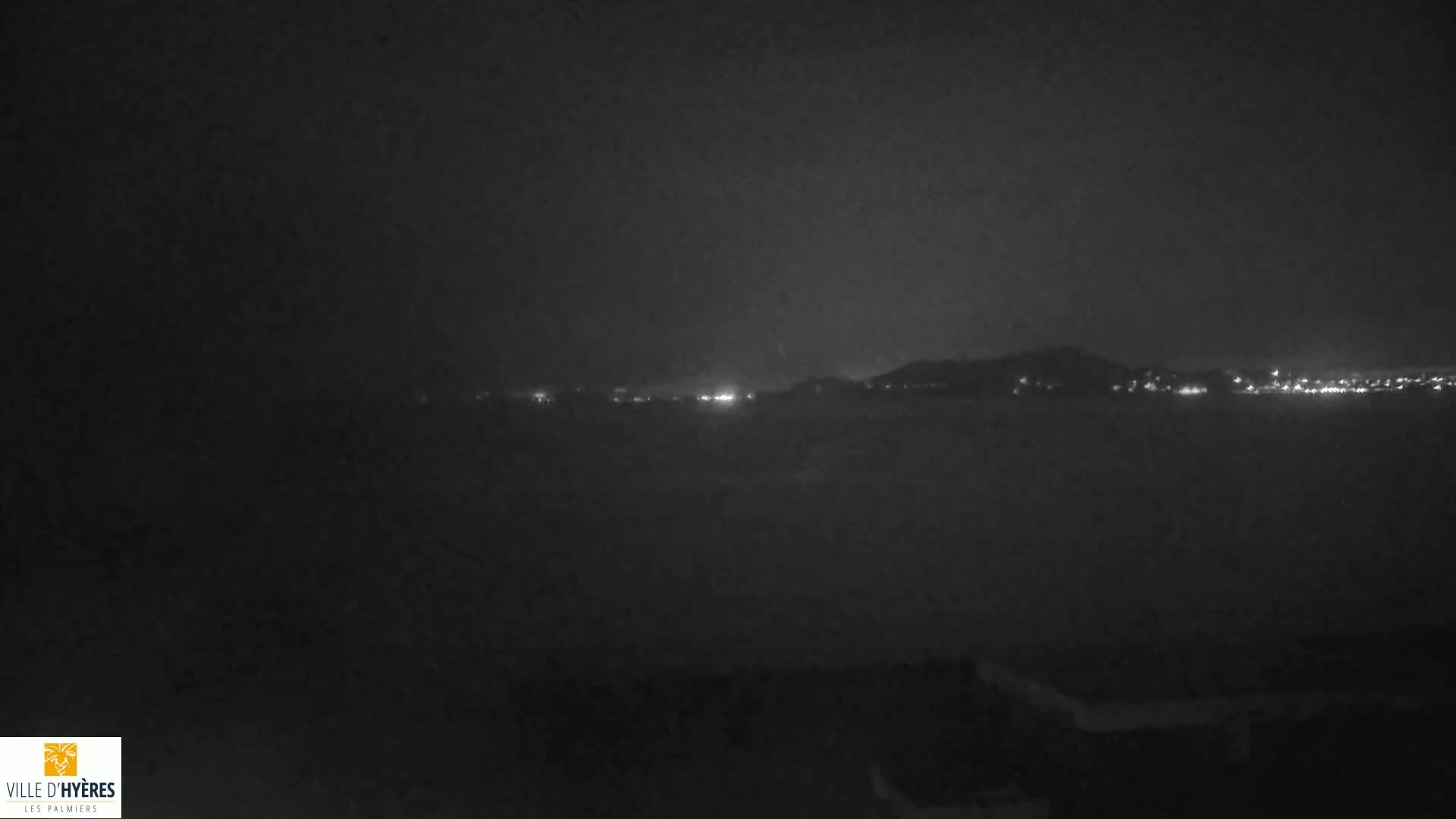 A nighttime long shot shows a dimly lit cityscape across a body of water under a dark, cloudy sky.