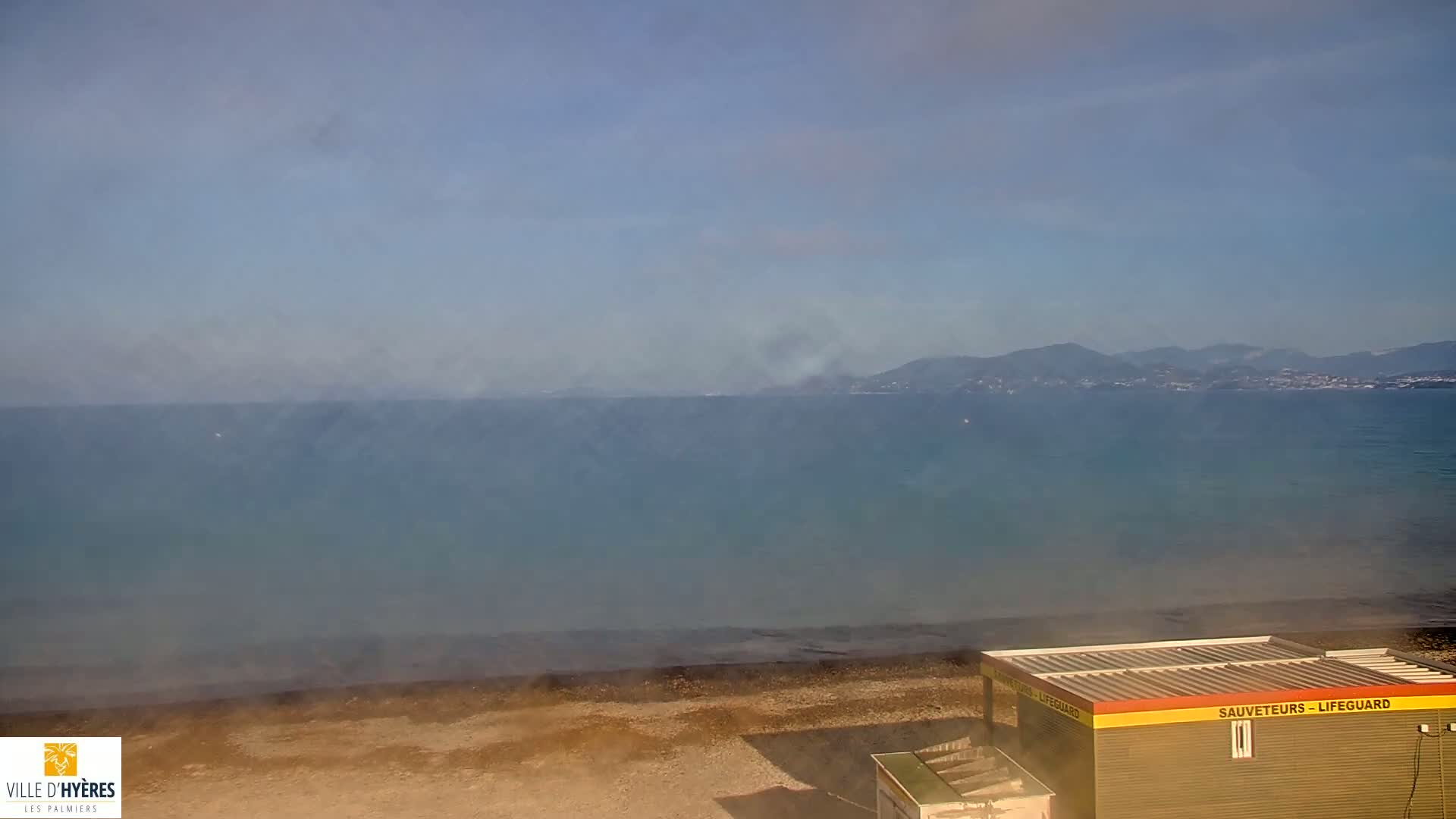 A hazy view shows a calm sea stretching to distant mountains, a sandy beach, and a small lifeguard station.