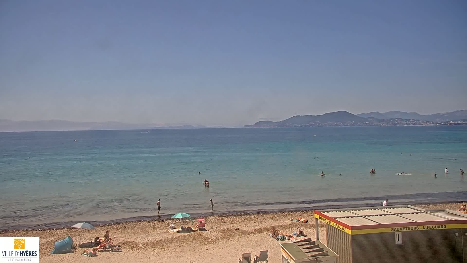 A sandy beach with scattered bathers and a lifeguard station overlooks a calm, turquoise sea under a clear blue sky, with distant mountains visible on the horizon.