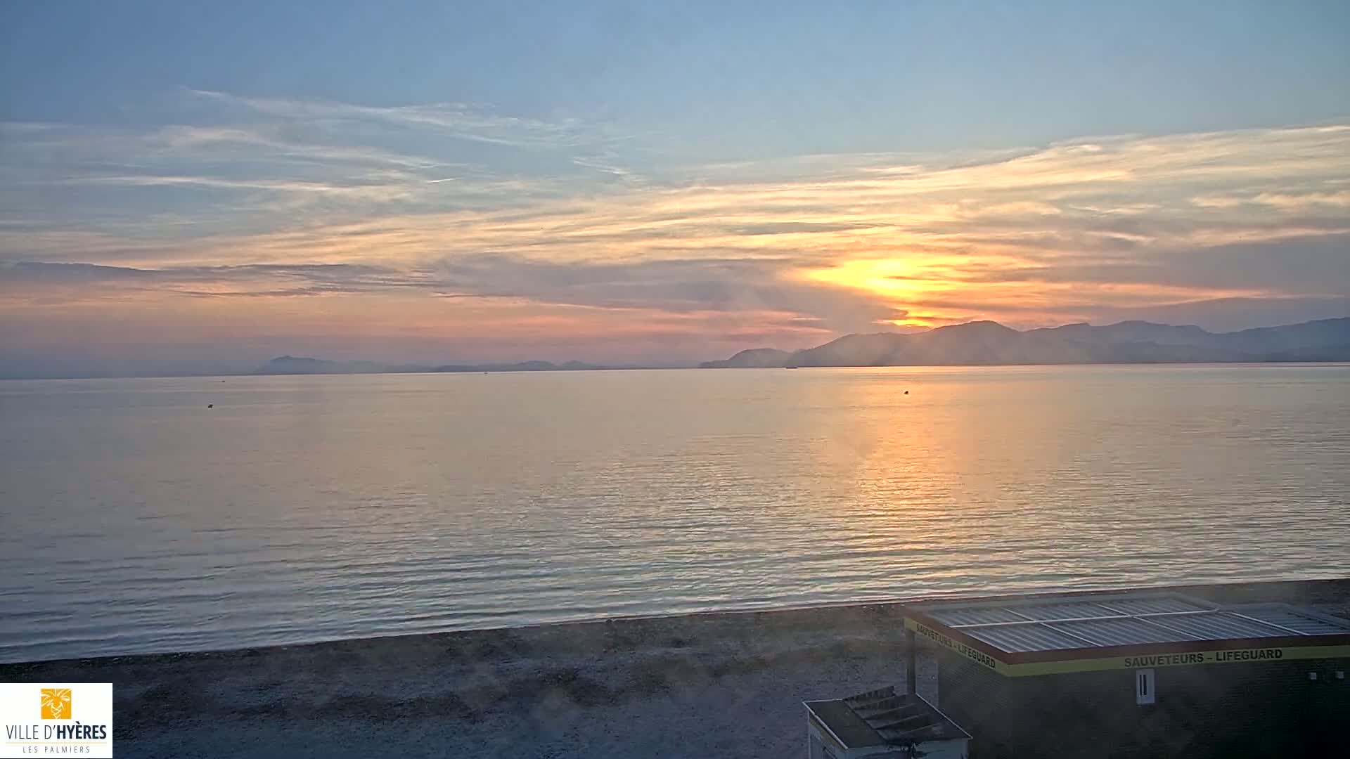 A calm sea at sunset with a lifeguard station on a sandy beach and mountains in the distance under a partly cloudy sky.