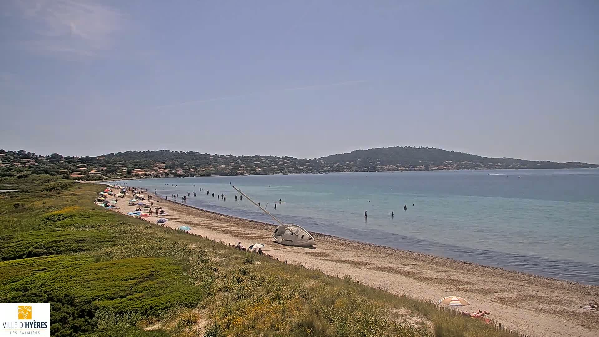 A sandy beach with many people sunbathing and swimming in calm, clear turquoise water, backed by green hills under a mostly clear sky.