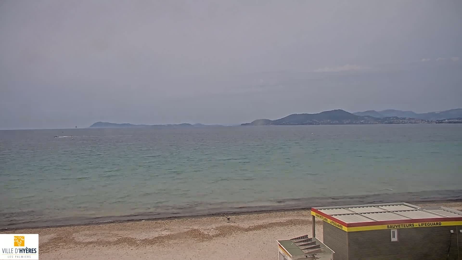 A lifeguard station sits on a sandy beach overlooking a calm, light-green sea and distant mountains under an overcast sky.