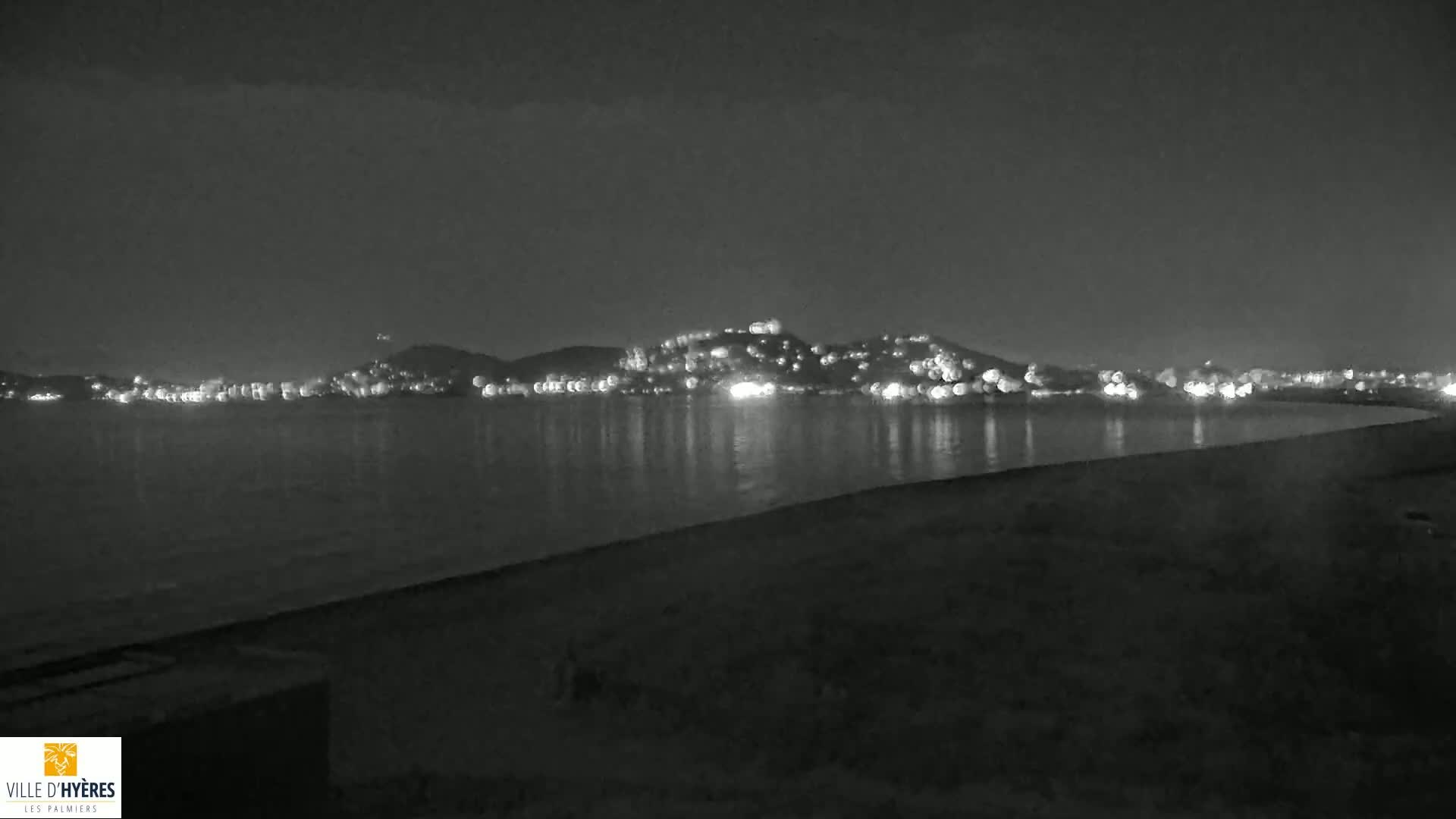 A nighttime view shows a hill dotted with lights across a calm body of water from a dark shoreline.