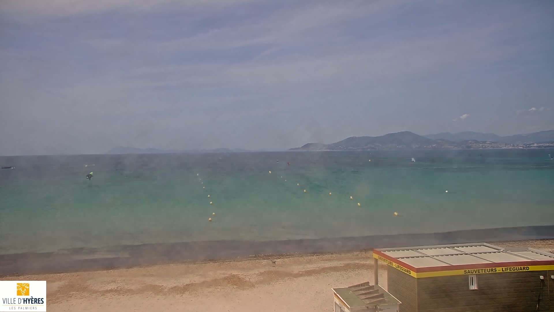A sandy beach with a lifeguard station overlooks a calm, teal-green ocean with a hazy atmosphere and distant mountains under a mostly cloudy sky.