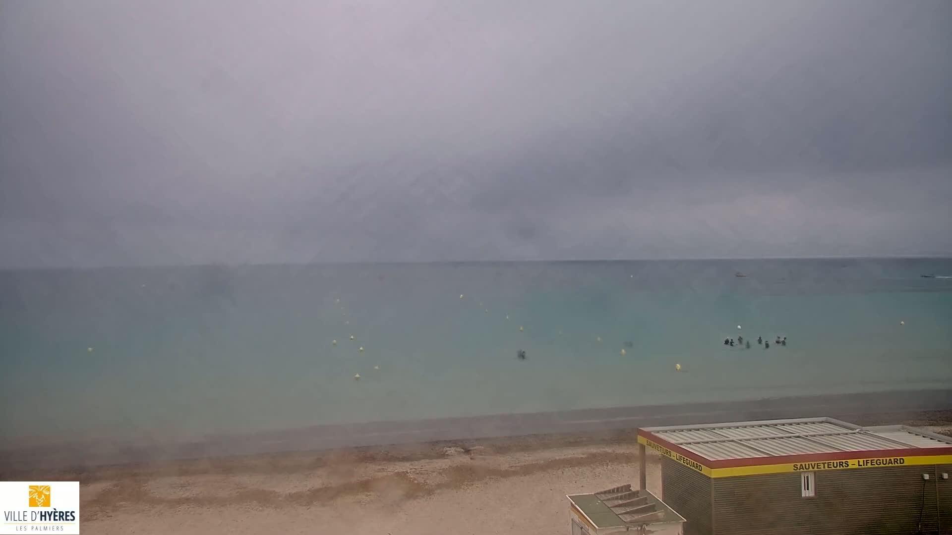 A lifeguard stand sits on a sandy beach overlooking a calm, light-blue sea under an overcast sky.