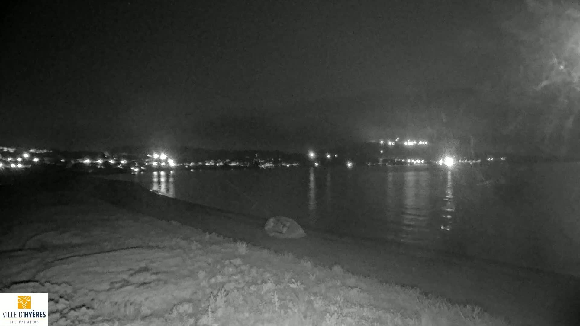A nighttime, low-light view shows a dark body of water reflecting distant lights across its surface, with a dark shoreline in the foreground.
