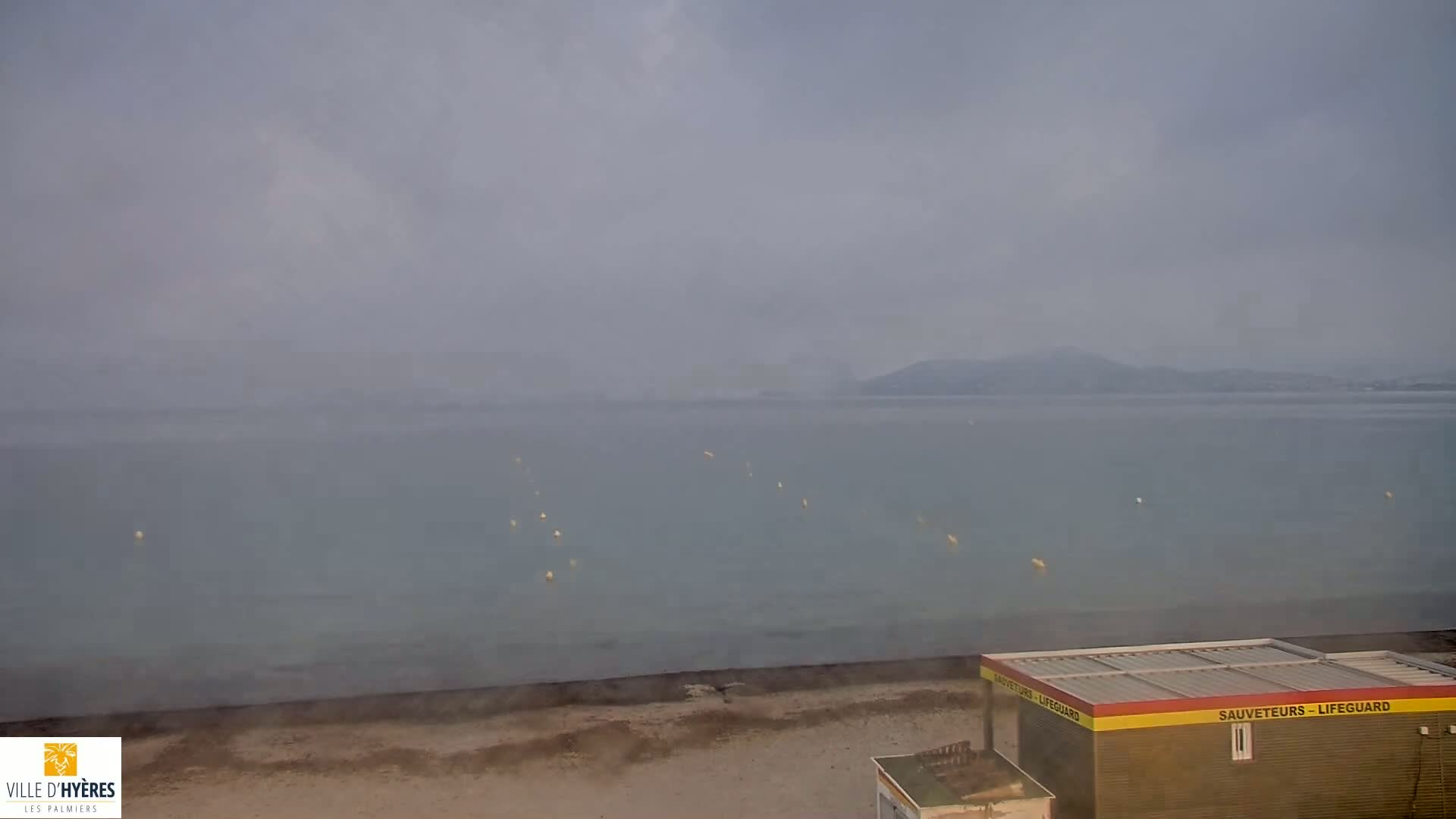 A hazy, overcast day reveals a calm body of water with a lifeguard station on a sandy beach and distant hills.