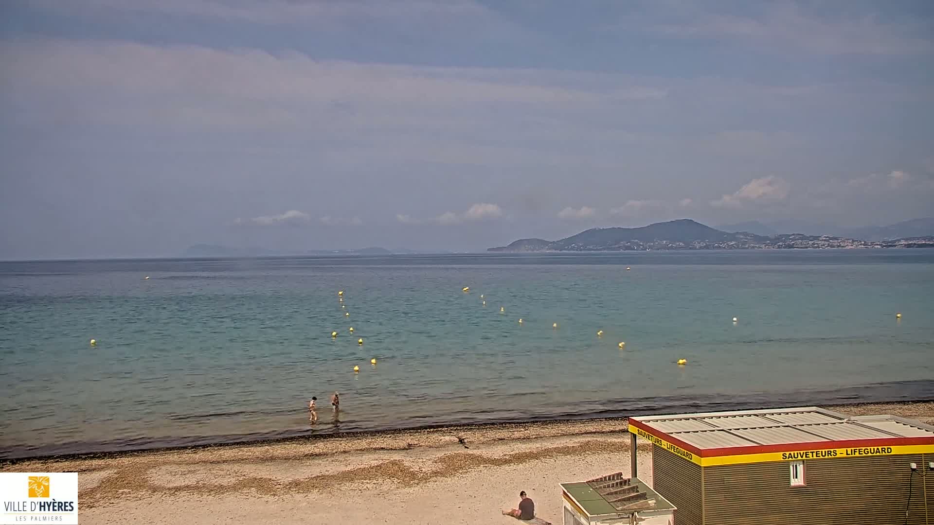 A calm, partly cloudy day at a beach shows a lifeguard station, a few people wading in the shallows, and a distant mountainous coastline.