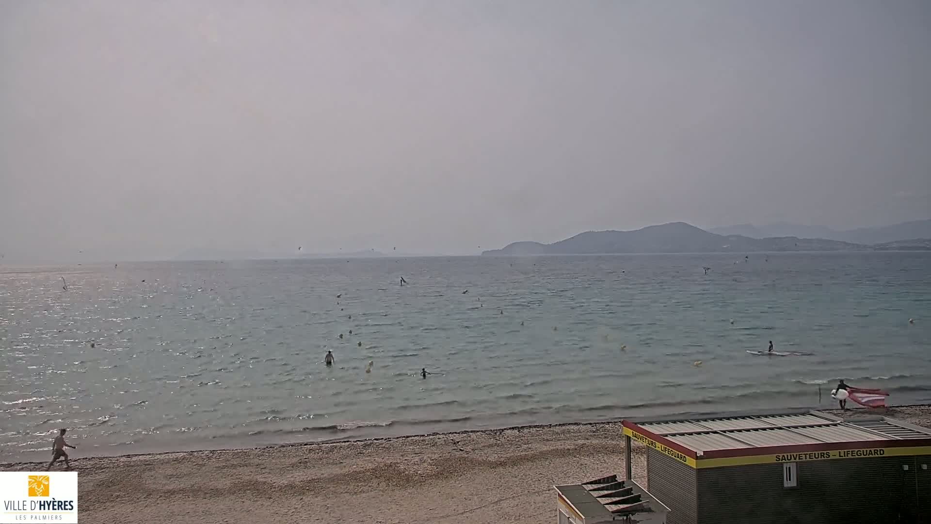 A hazy day at a beach shows people swimming and windsurfing in calm water, with distant mountains visible under a light-grey sky.