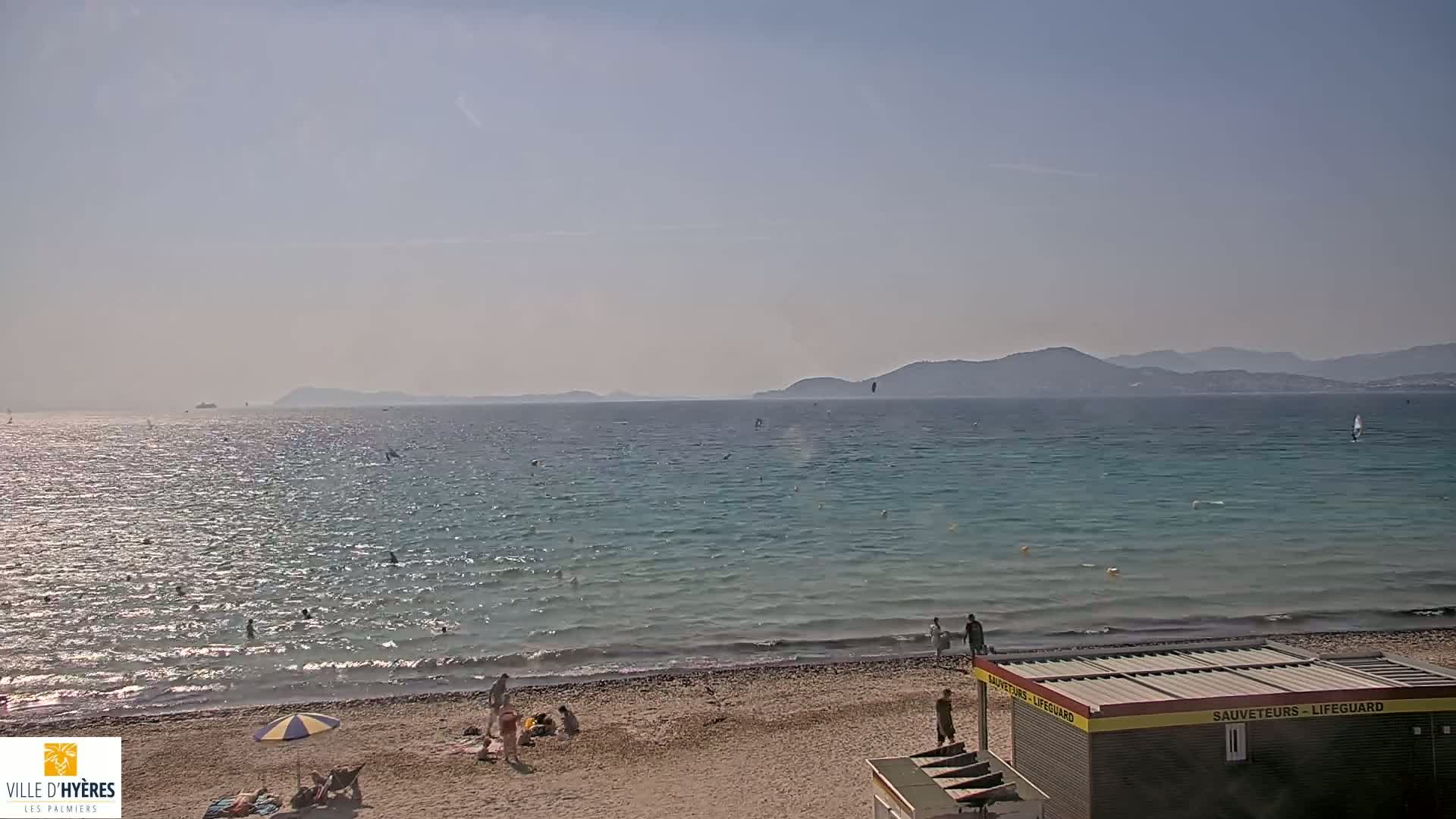 A sunny day at a beach with calm, light blue water, a few people swimming and sunbathing, and distant mountains visible on the horizon.