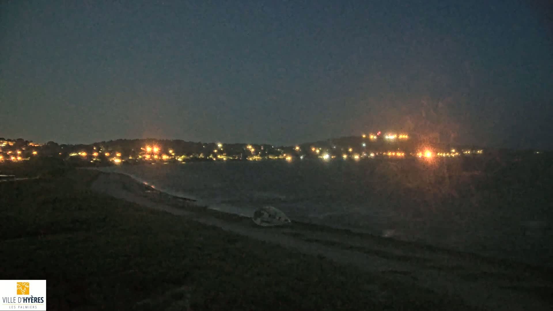 A dark, nighttime scene shows a distant hilltop illuminated with scattered lights across a dark body of water, under a clear, night sky.