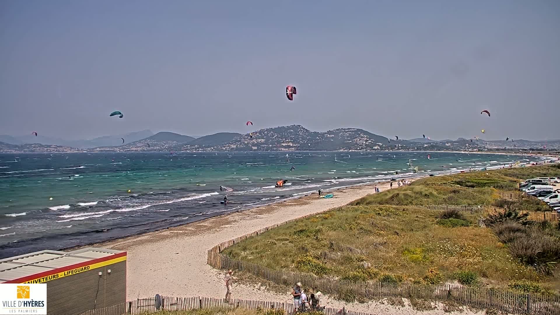 A sandy beach with several kitesurfers in the water and more kites visible in the air, under a sunny sky with a hazy distant mountain range in the background.