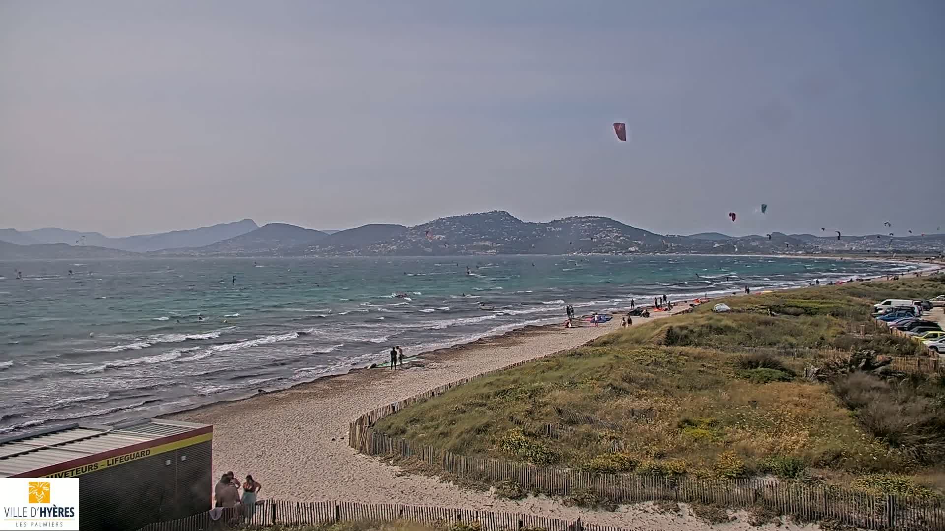 A sandy beach with several people and numerous kitesurfers in the water, under a hazy sky, backed by distant mountains.