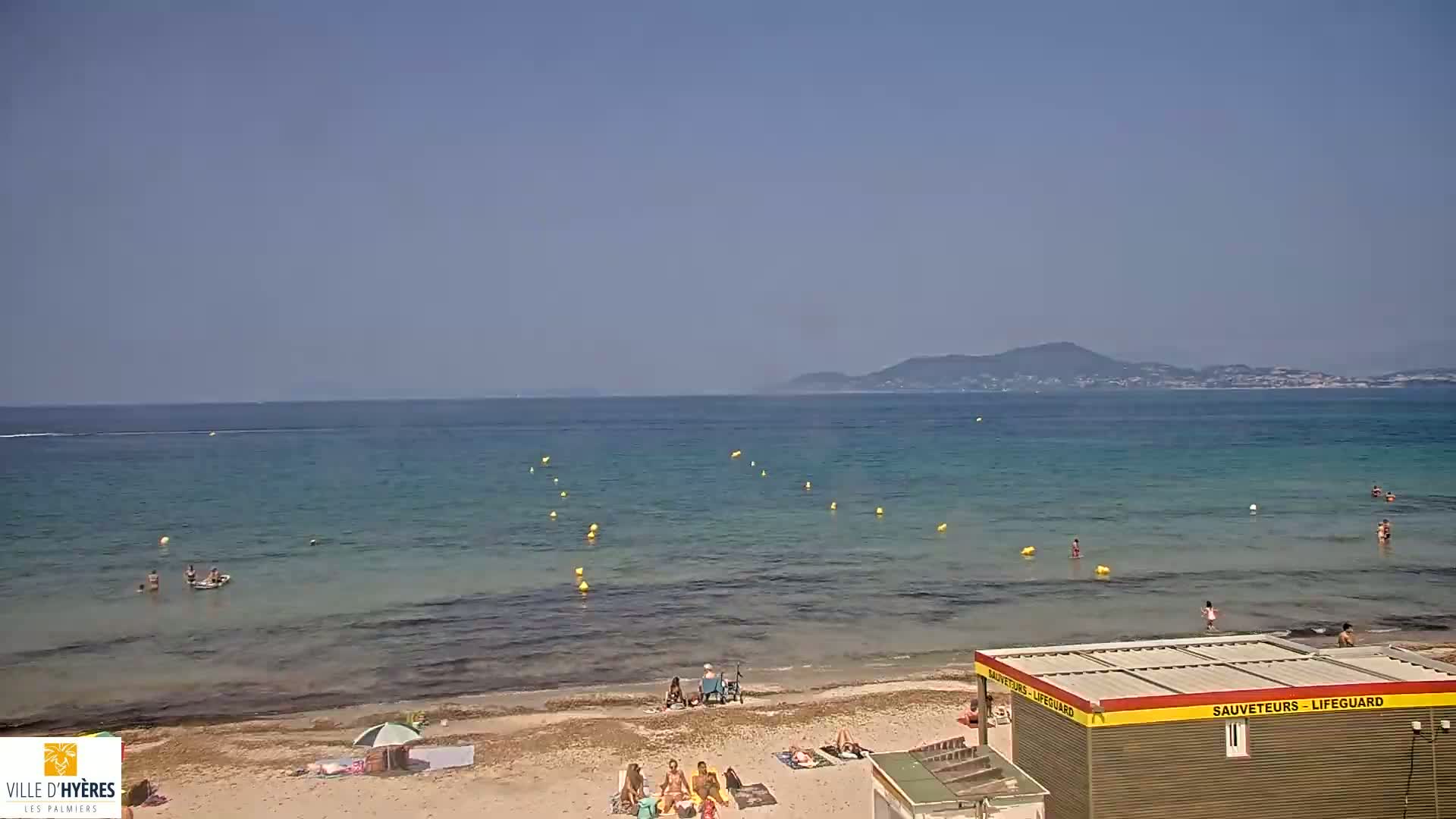 A sandy beach with a few people sunbathing and swimming in a calm, clear sea under a clear blue sky, with a distant mountain range visible on the horizon.