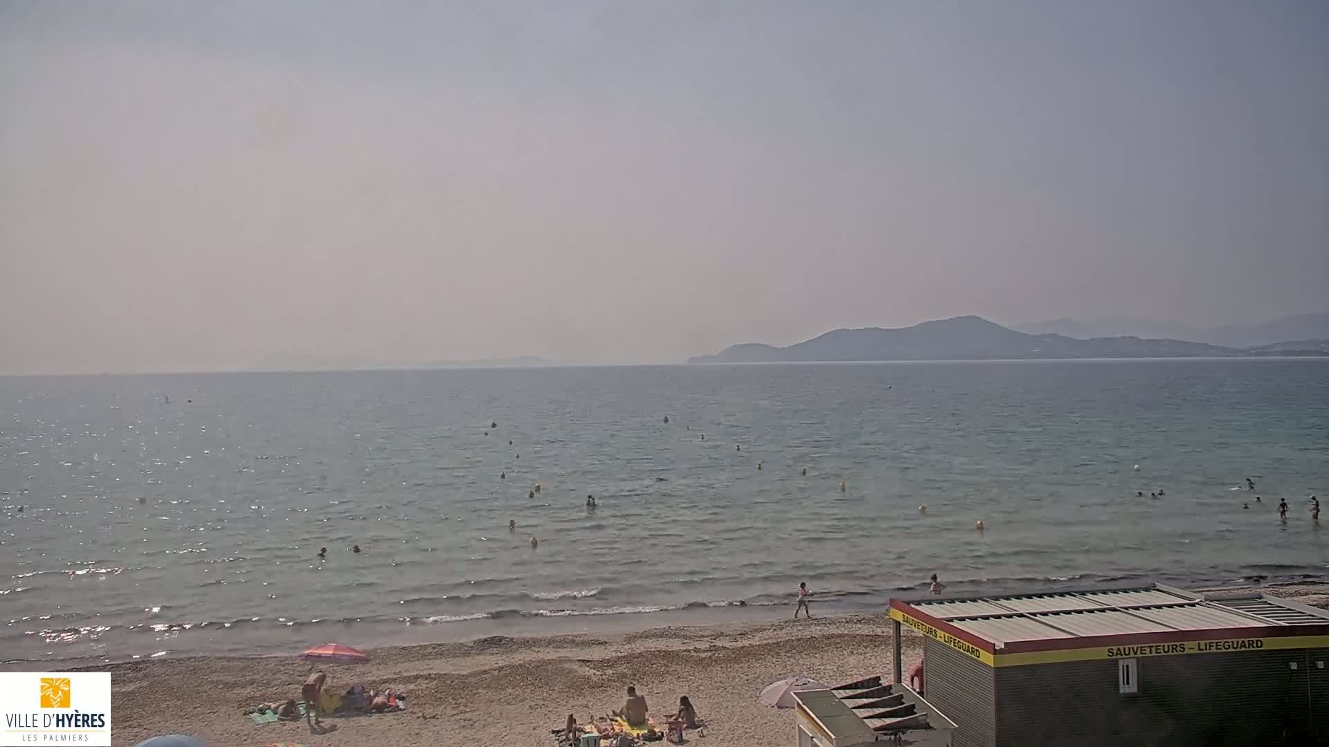 A hazy, sunny day at a beach shows calm, clear water with people swimming and sunbathing on the sand near a lifeguard station and distant mountains.