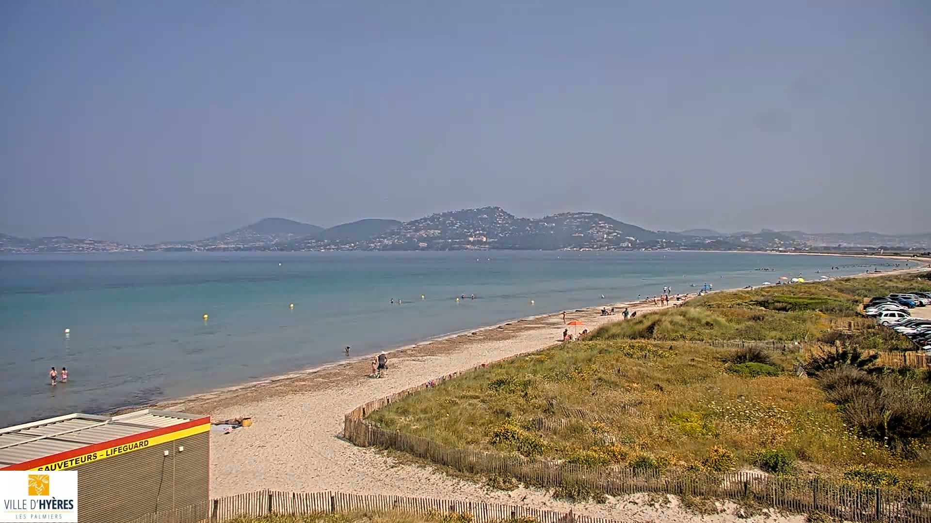 A sandy beach with calm, turquoise water stretches toward a distant mountain range under a clear, sunny sky.