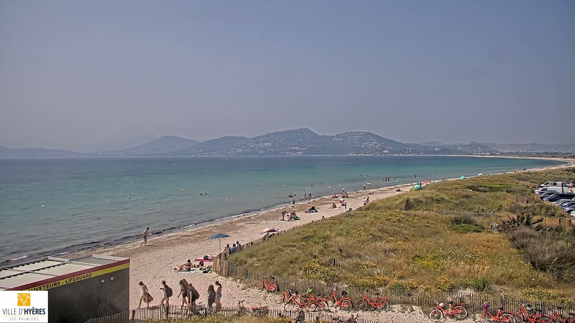 A sandy beach with people sunbathing and swimming, backed by grassy dunes, overlooks a calm, turquoise sea and distant mountains under a hazy sky.