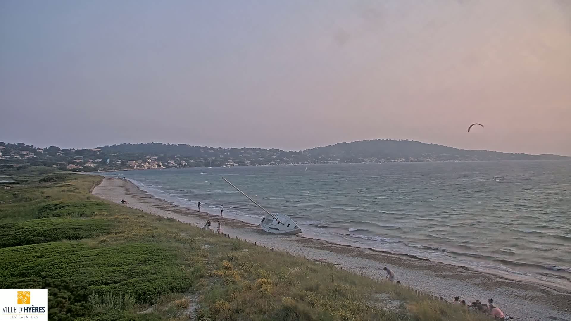 A sandy beach with a capsized sailboat, several people, and a kite surfer are visible against the backdrop of a calm sea and a distant coastline under a dusky sky.