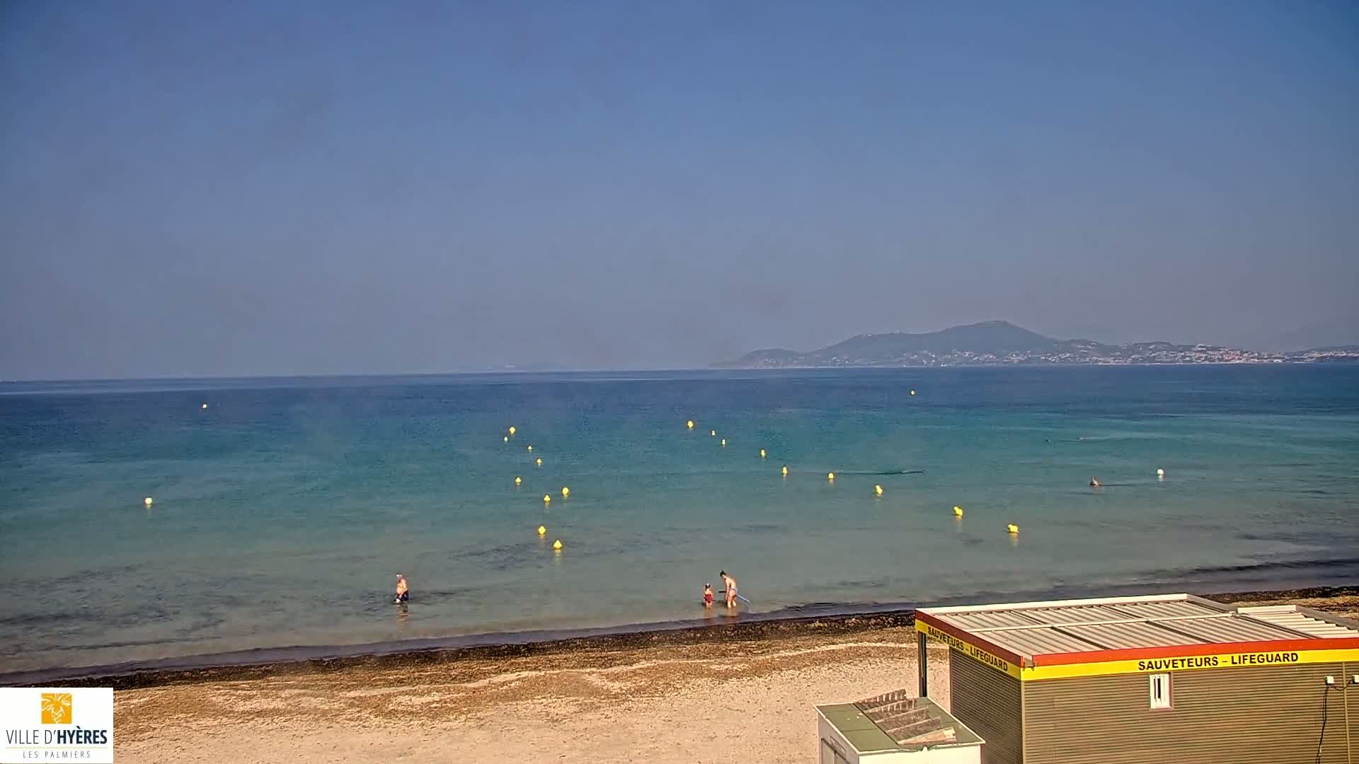 A sandy beach with calm, clear turquoise water, a lifeguard station, and a few people swimming or wading, under a clear blue sky.