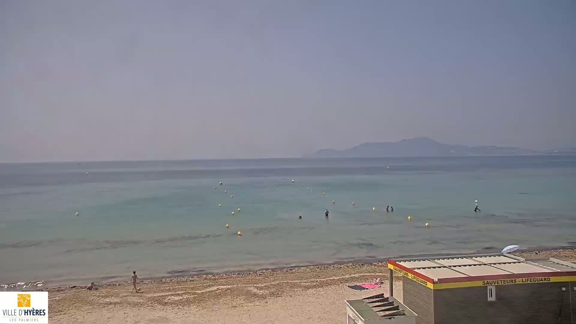 A lifeguard station sits on a sandy beach overlooking calm, shallow turquoise water with several people swimming and a hazy mountain range in the distance under a clear sky.
