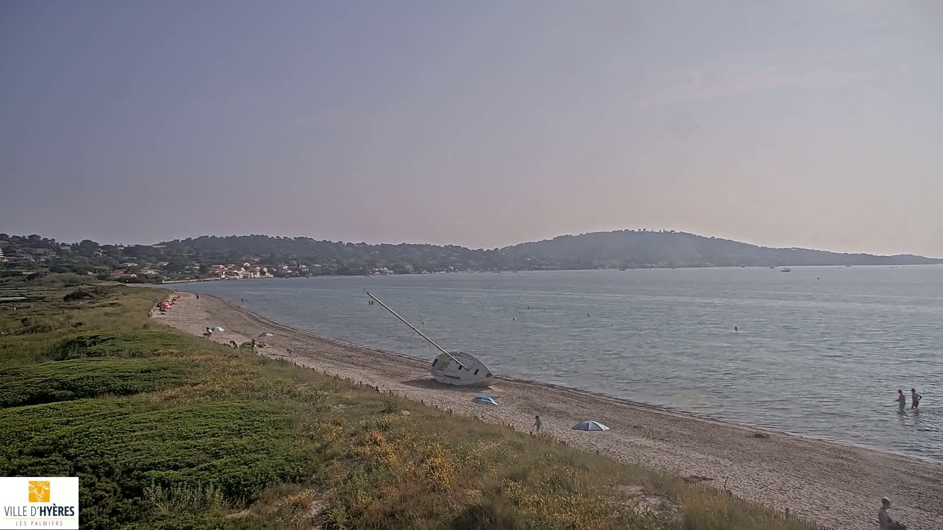 A sandy beach with a capsized sailboat on the shore, a calm sea, and a few people are visible under a clear sky.