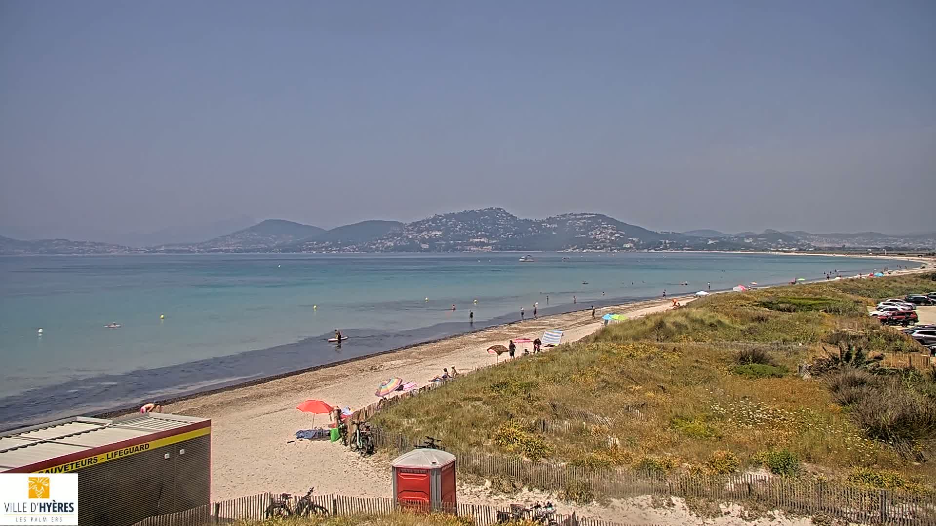 A sandy beach with turquoise water and scattered bathers stretches along a coastline, backed by hills under a clear, sunny sky.