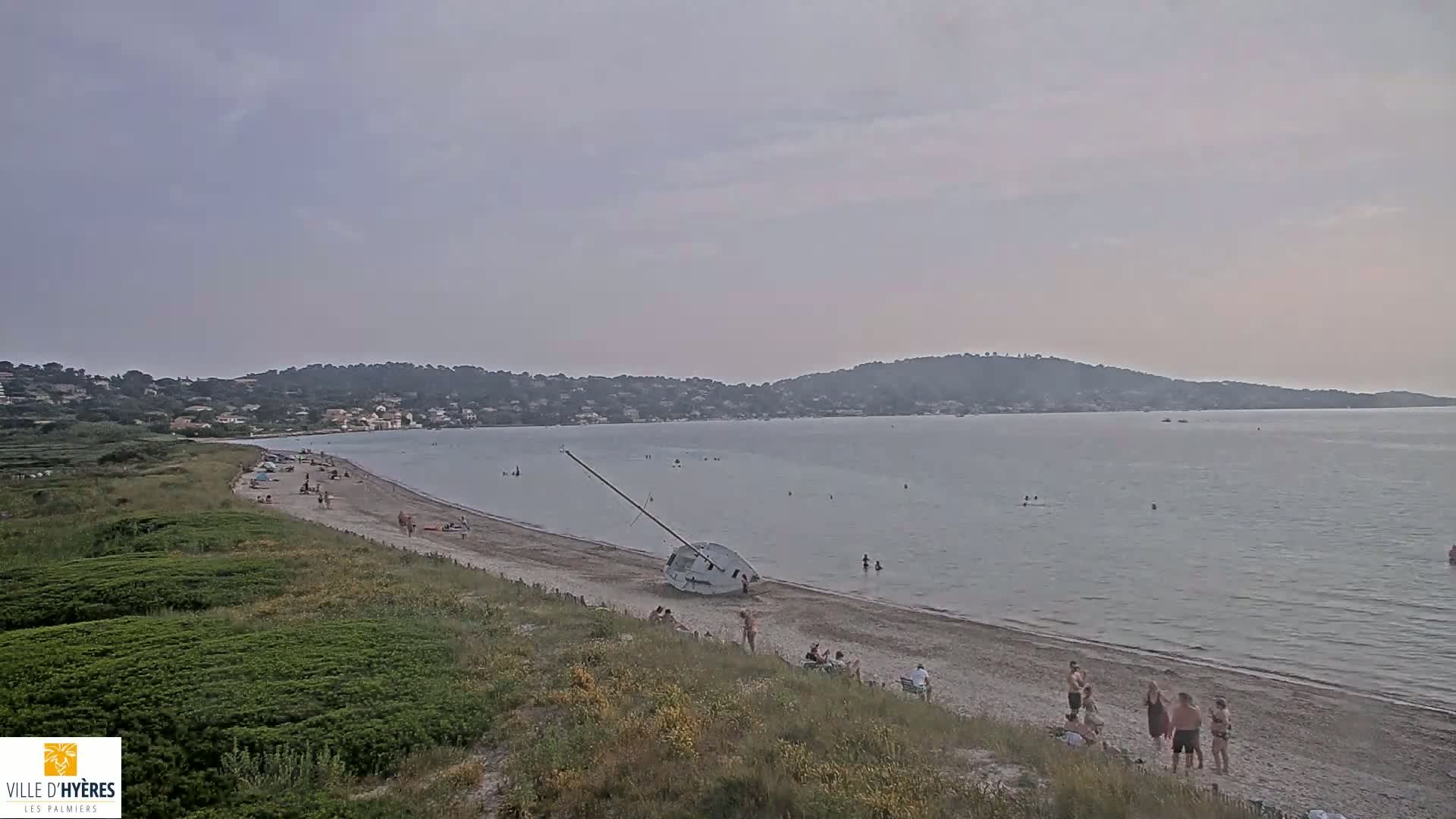 A sandy beach with scattered people, a capsized sailboat, and calm, light-grey water under an overcast sky, is backed by low green hills and a hazy line of distant buildings.