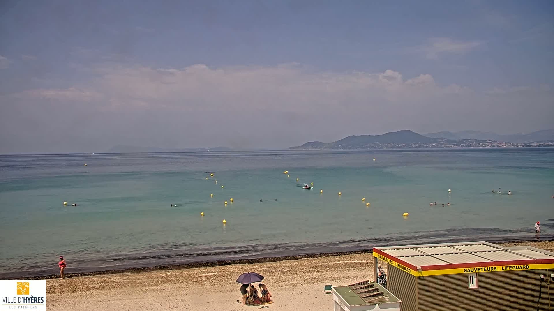 A sandy beach with calm, clear turquoise water, a few people swimming and sunbathing, and a lifeguard station under a partly cloudy sky with distant mountains visible on the horizon.