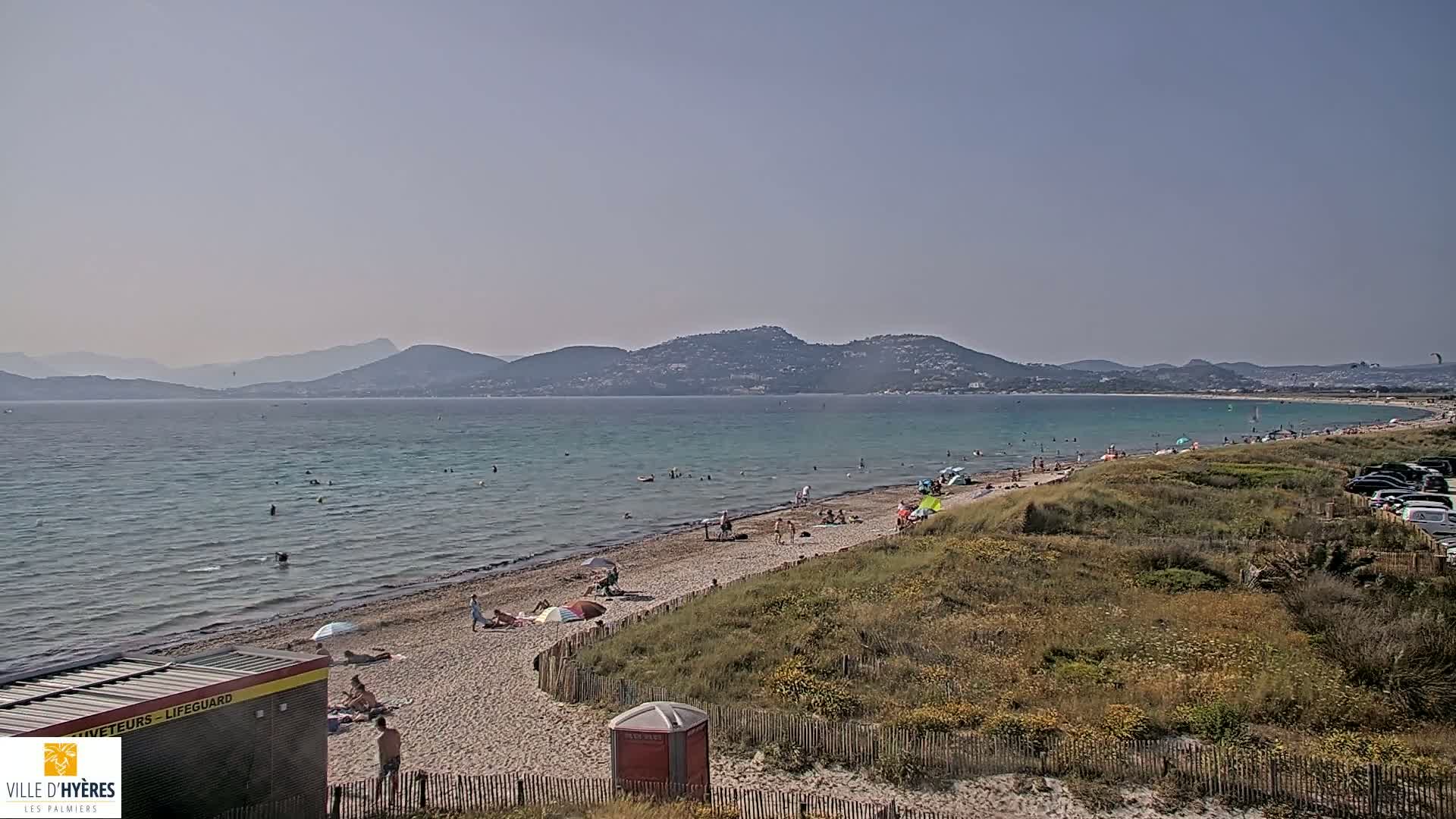 A sandy beach with scattered bathers stretches along a calm, turquoise sea, backed by a line of distant mountains under a hazy sky.