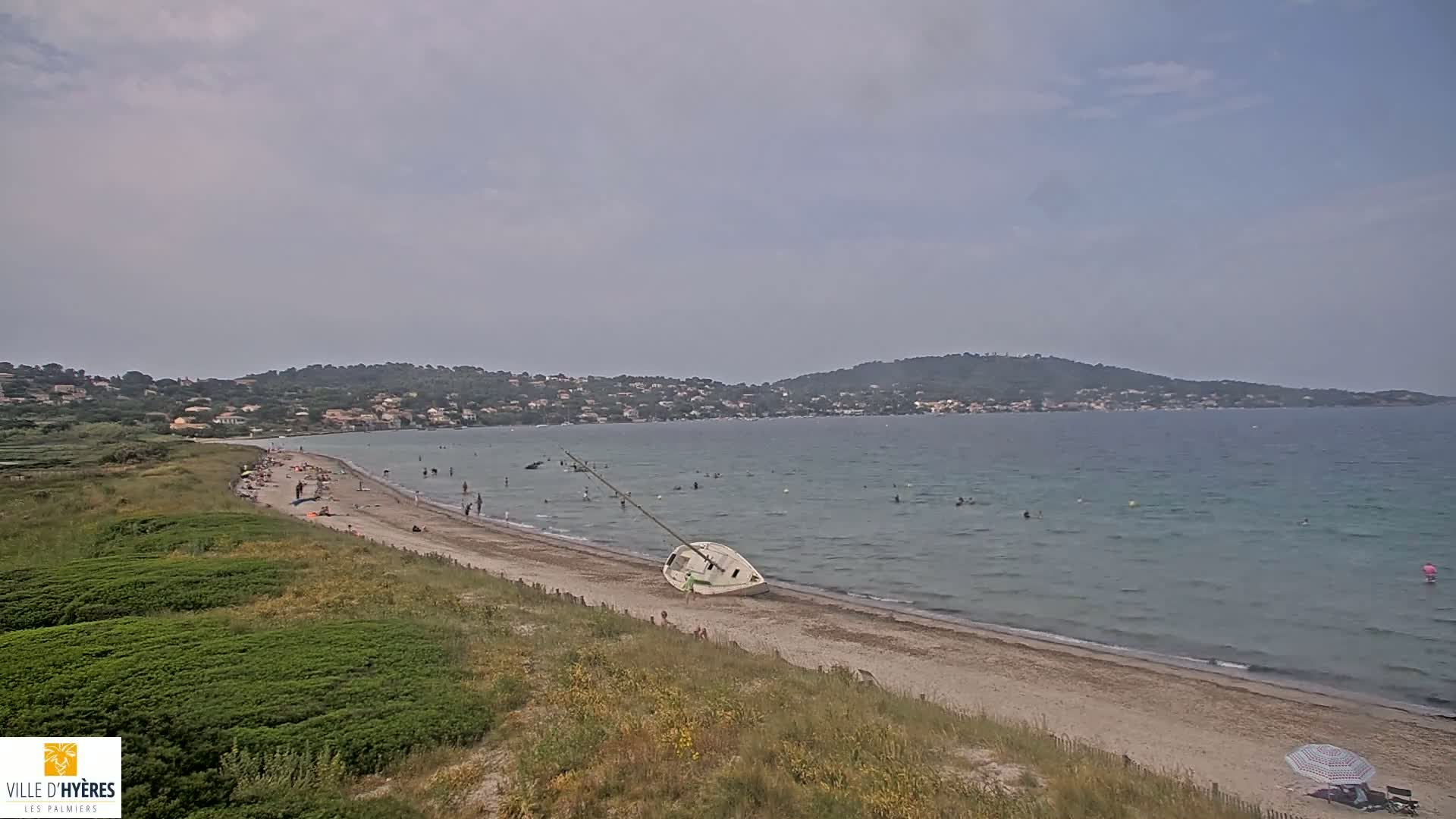 A sandy beach with scattered swimmers and sunbathers, a capsized sailboat, and green vegetation overlooking a calm sea under an overcast sky.
