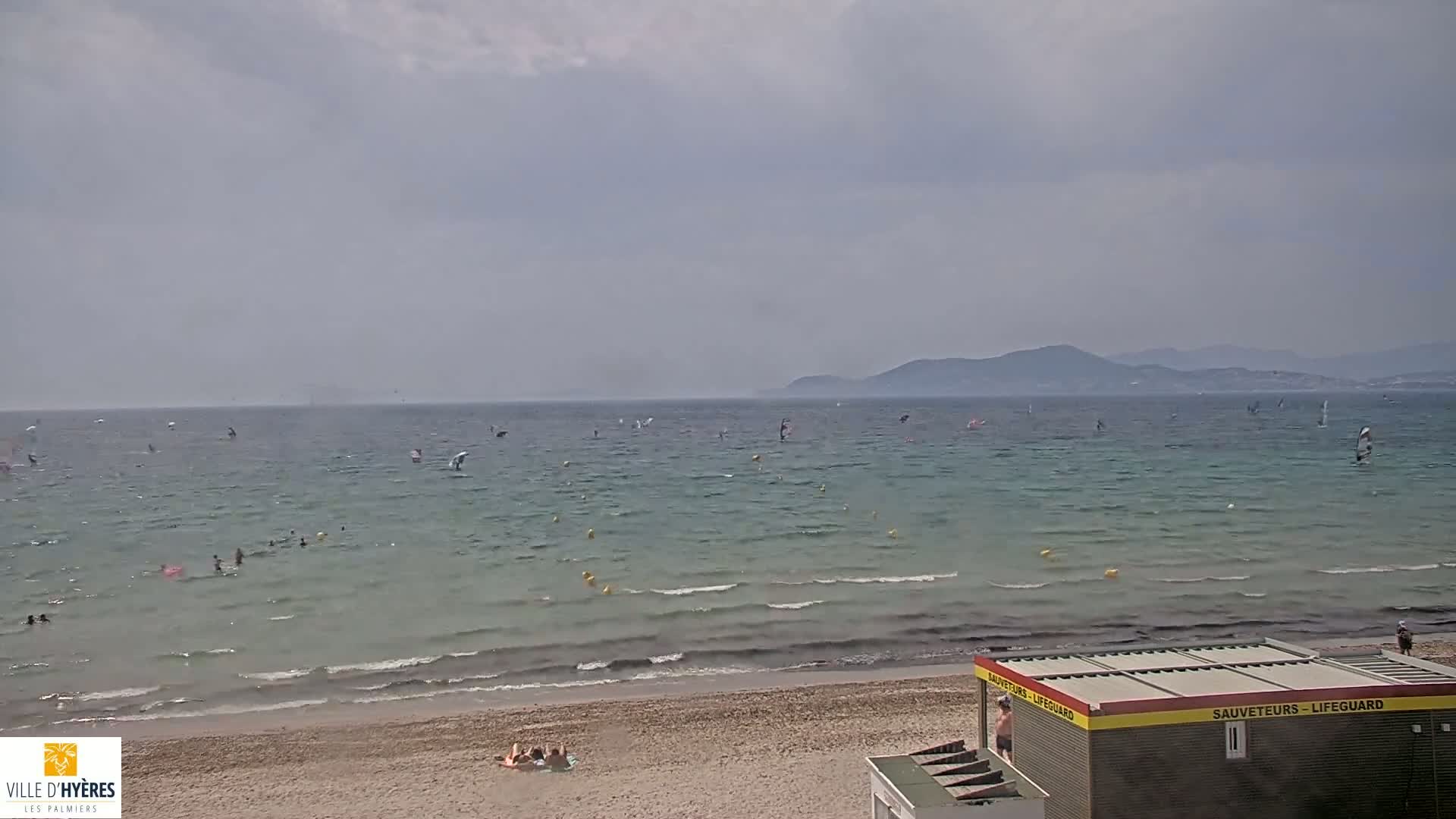 A sandy beach with calm, light-blue water shows several windsurfers in the distance under a mostly cloudy sky, with a lifeguard stand visible on the shore.
