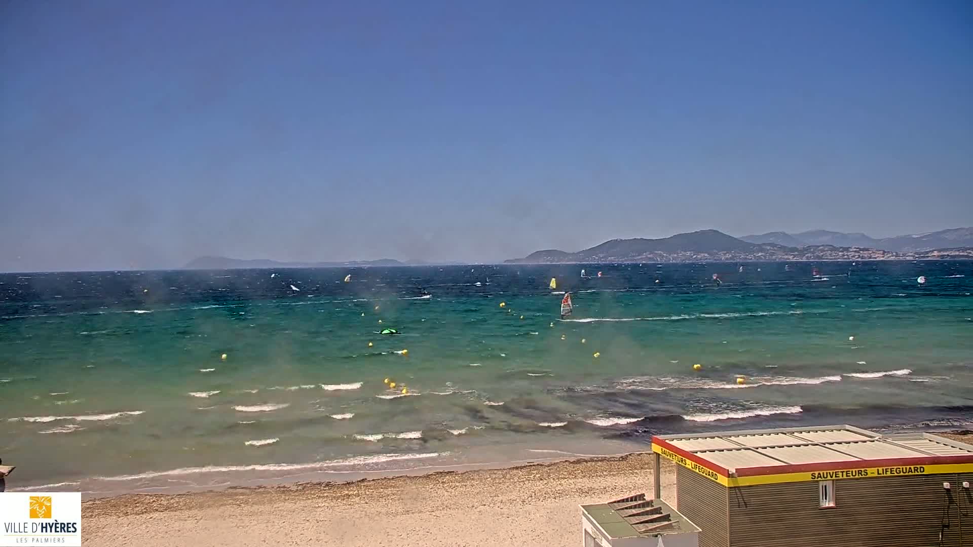 A windy, sunny day at a beach with numerous windsurfers on the water, mountains in the distance, and a lifeguard station on the shore.