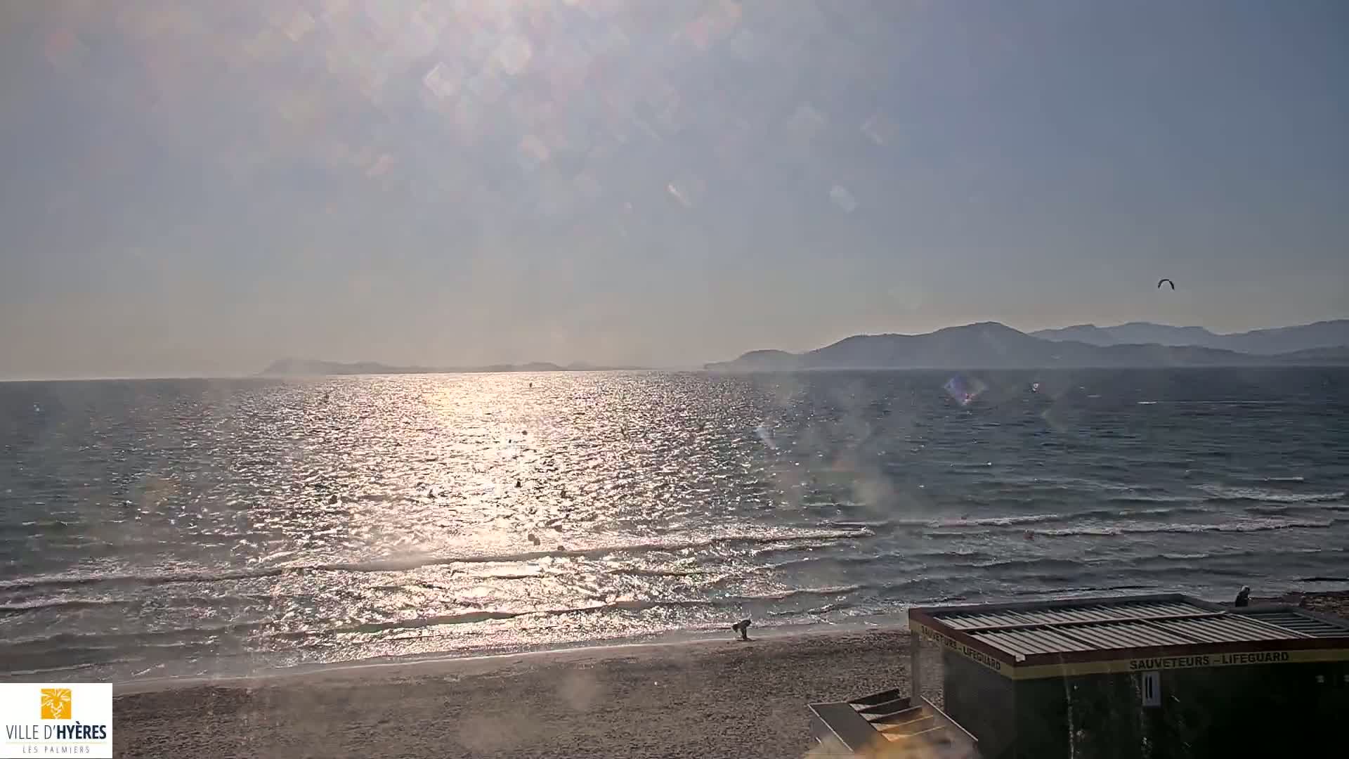 A sandy beach with calm, sunlit water stretches to a distant mountain range under a mostly clear sky, with a few people visible on the beach and in the water.