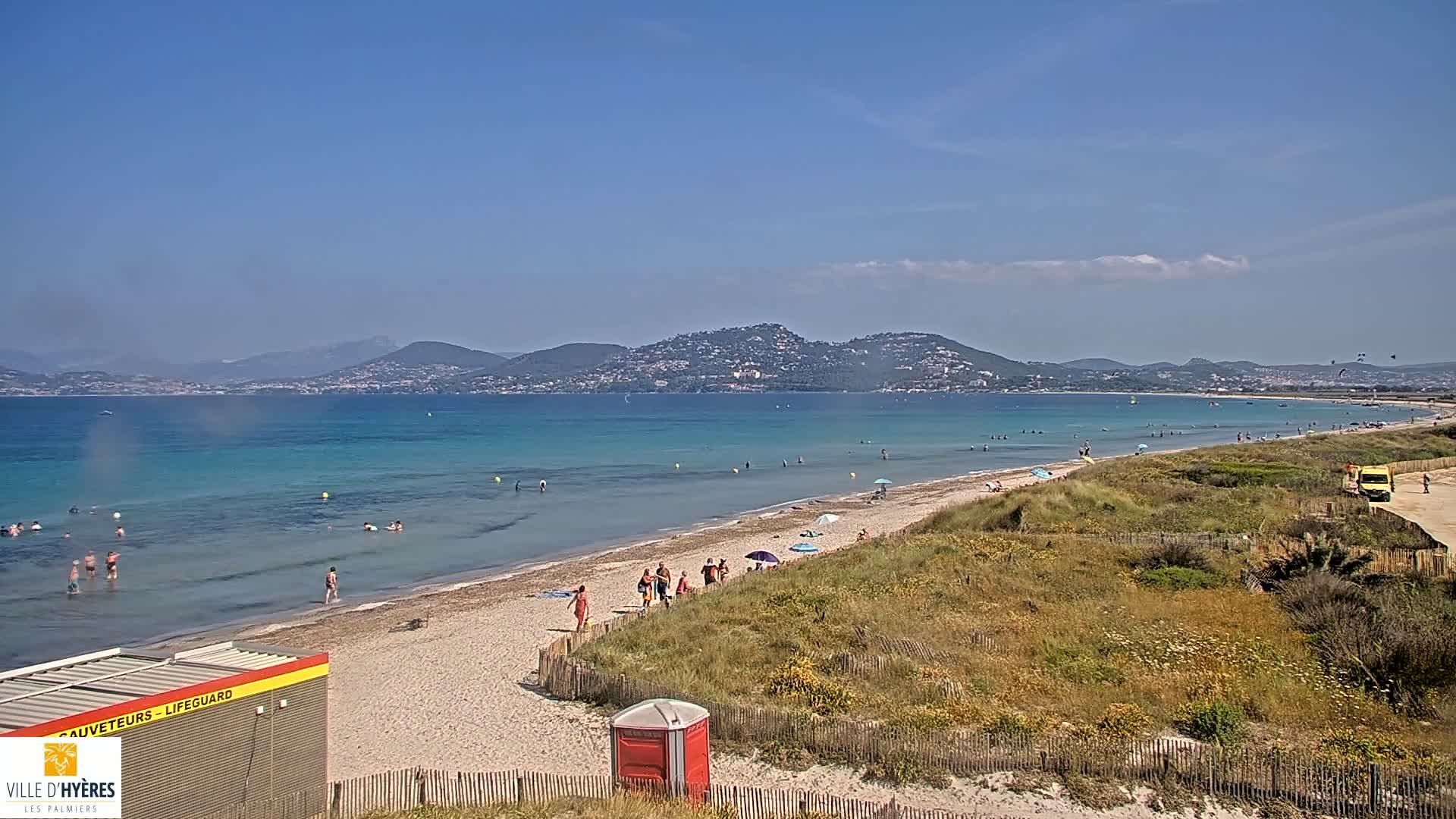 A sandy beach with turquoise water and scattered bathers stretches out before a line of distant mountains under a mostly clear sky.