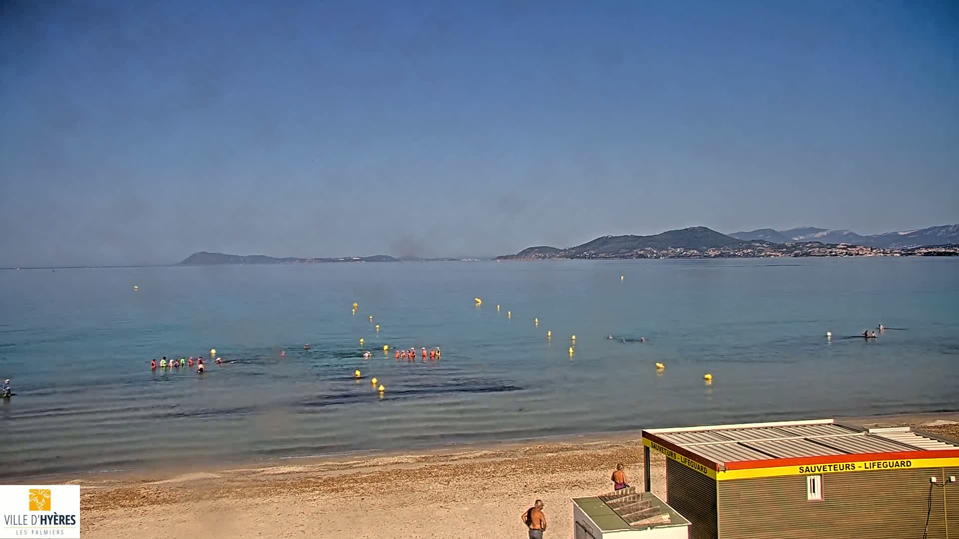 A sandy beach with people swimming in calm, clear water under a clear blue sky, with distant mountains in the background.