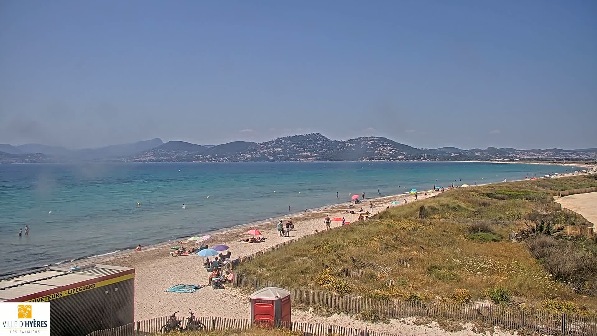 A sandy beach with scattered people under umbrellas stretches along a calm, turquoise ocean, backed by a line of low hills under a clear blue sky.