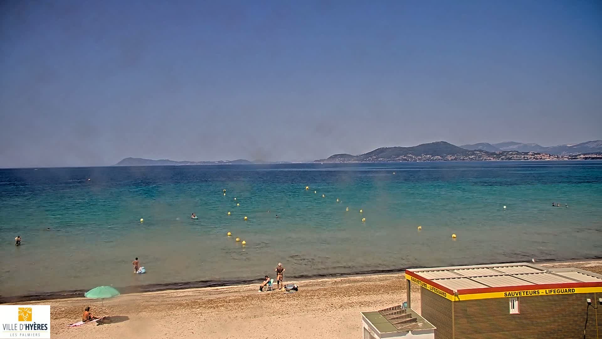 A sandy beach with a few people swimming and sunbathing, a lifeguard station, and calm, clear turquoise water extending to distant mountains under a clear blue sky.