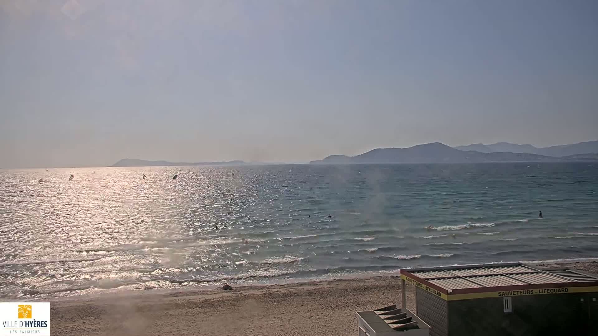 A sandy beach with calm, glittering water extends to a hazy mountainous coastline under a sunny sky, with several people swimming or windsurfing in the sea.