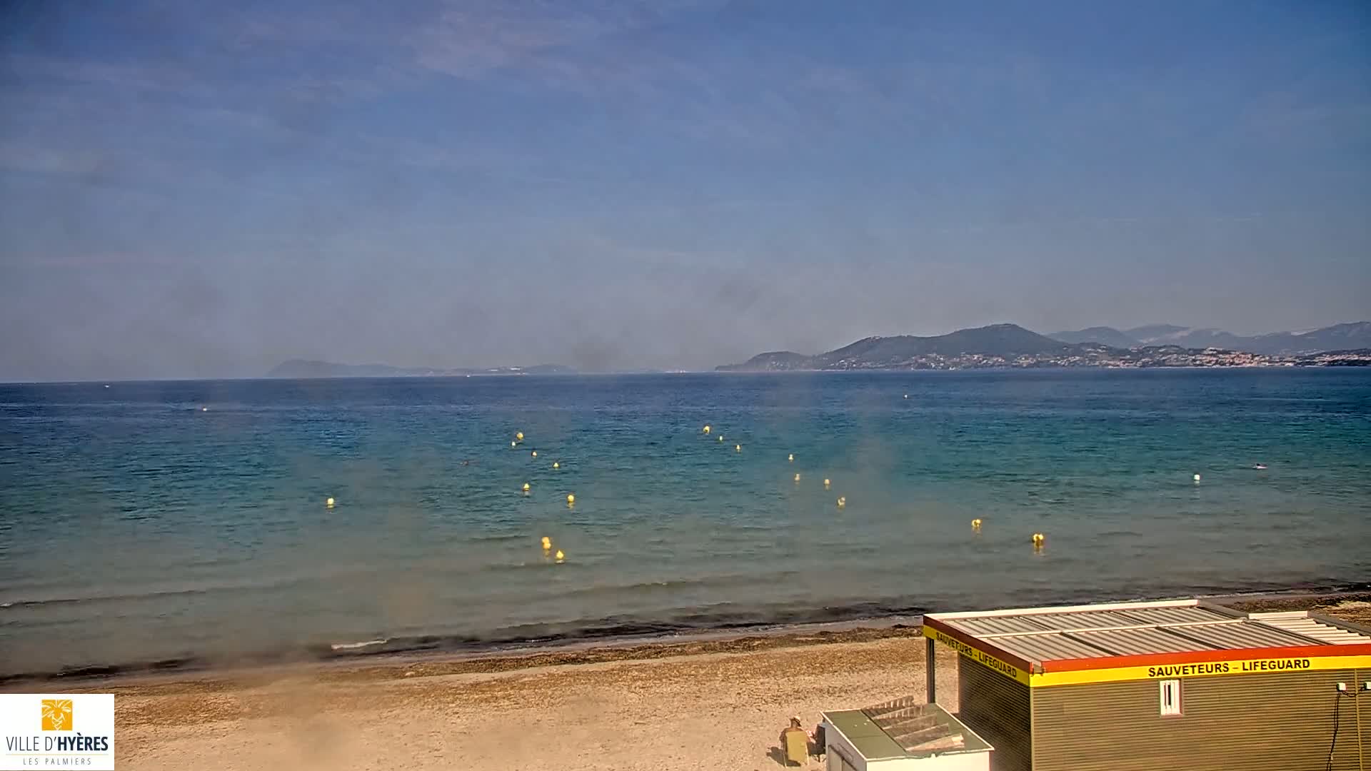 A sandy beach with calm, clear water, yellow buoys, and a lifeguard station is visible, with mountains in the background under a mostly clear sky.